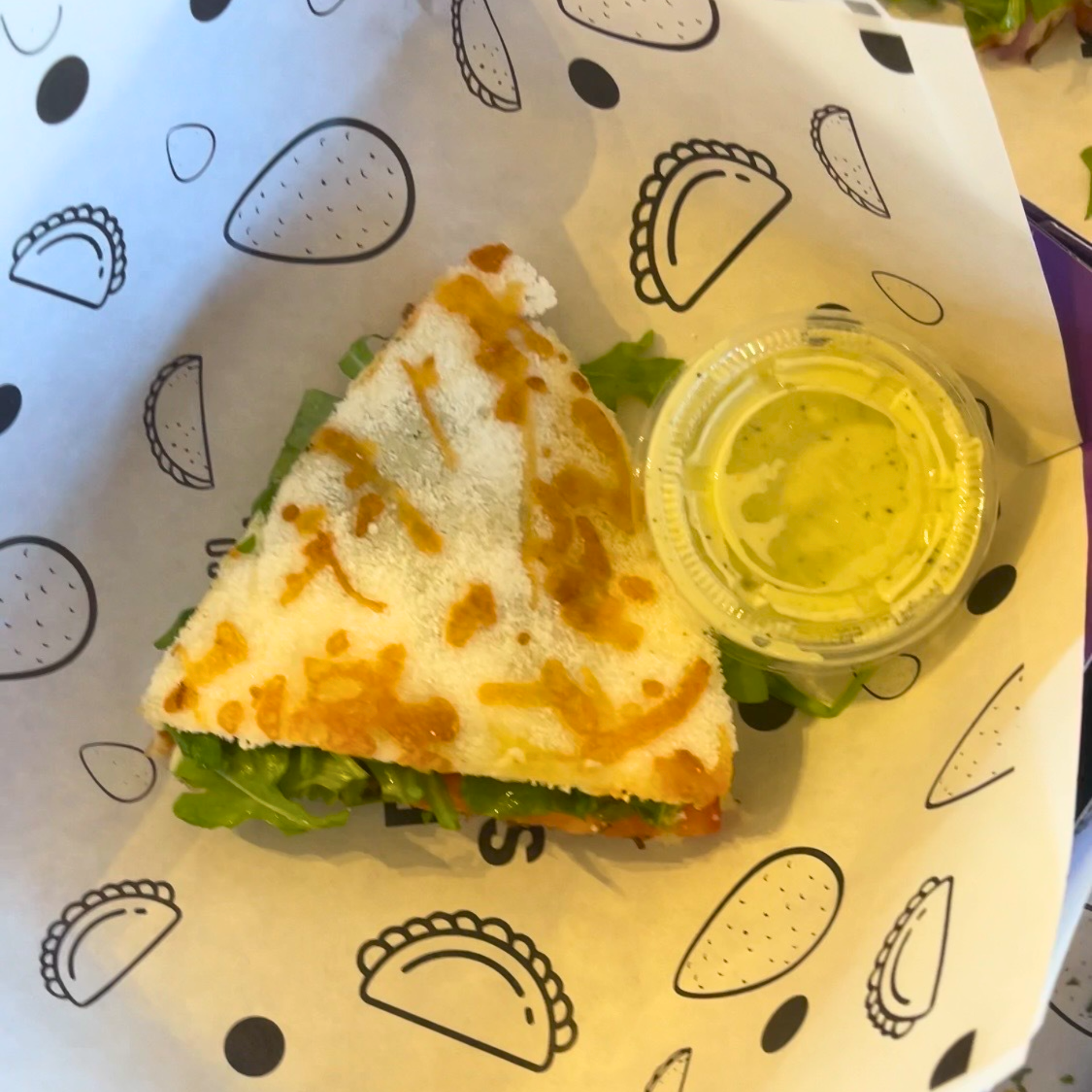 Toasted triangular sandwich with leafy greens, served on printed paper with a small cup of green sauce