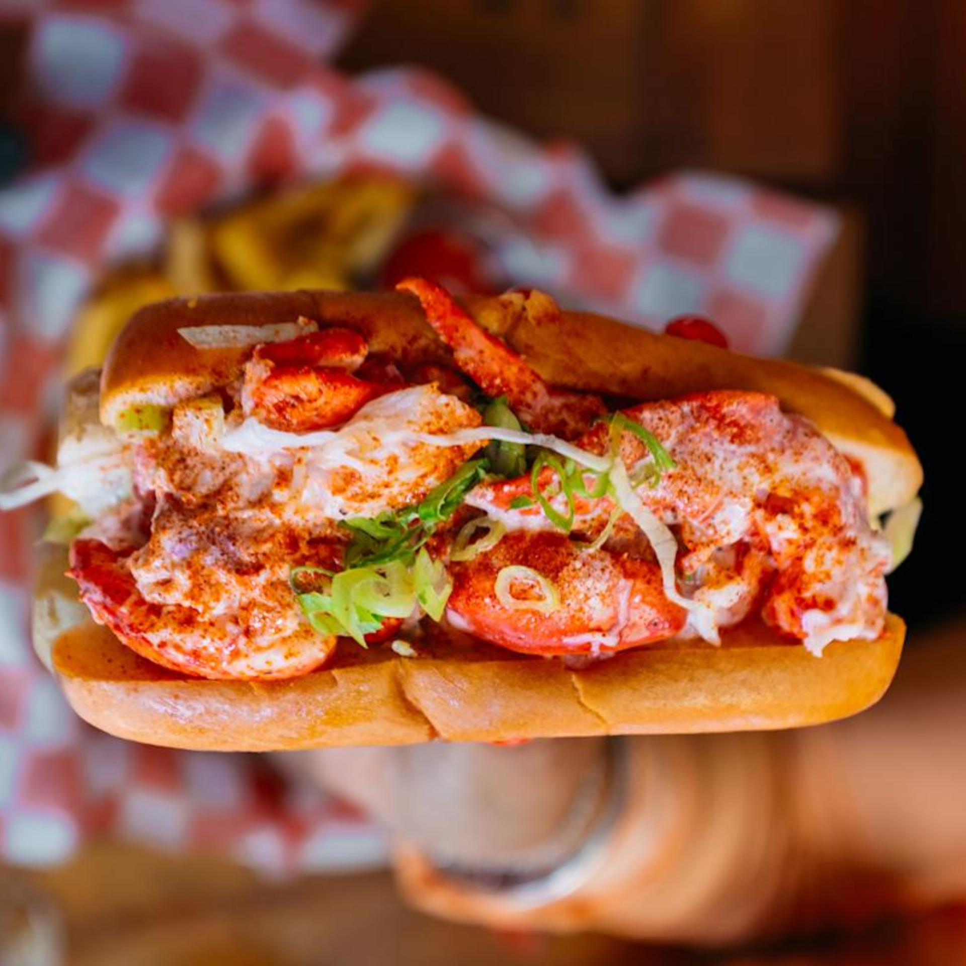 A close-up shot of a hand holding a New England-style lobster roll in a toasted, buttered split-top bun. The roll is filled with large chunks of lobster meat seasoned with paprika and topped with sliced green scallions, with a red and white checkered paper liner in the background