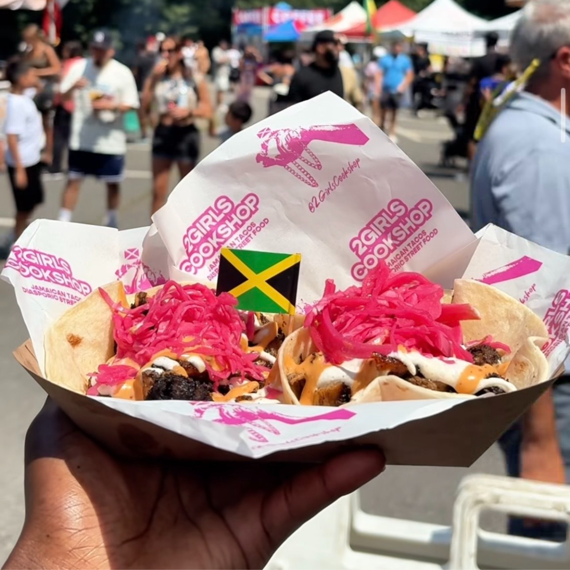 Tray of tacos topped with pink pickled onions, sauces, grilled meat, and a small Jamaican flag