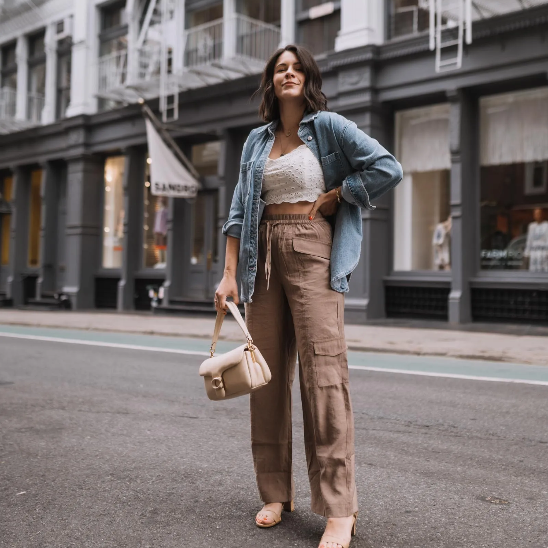 A woman in a denim shirt, white lace crop top, and tan cargo pants stands on a New York City street. She holds a cream-colored handbag while posing in front of grey buildings with white storefronts