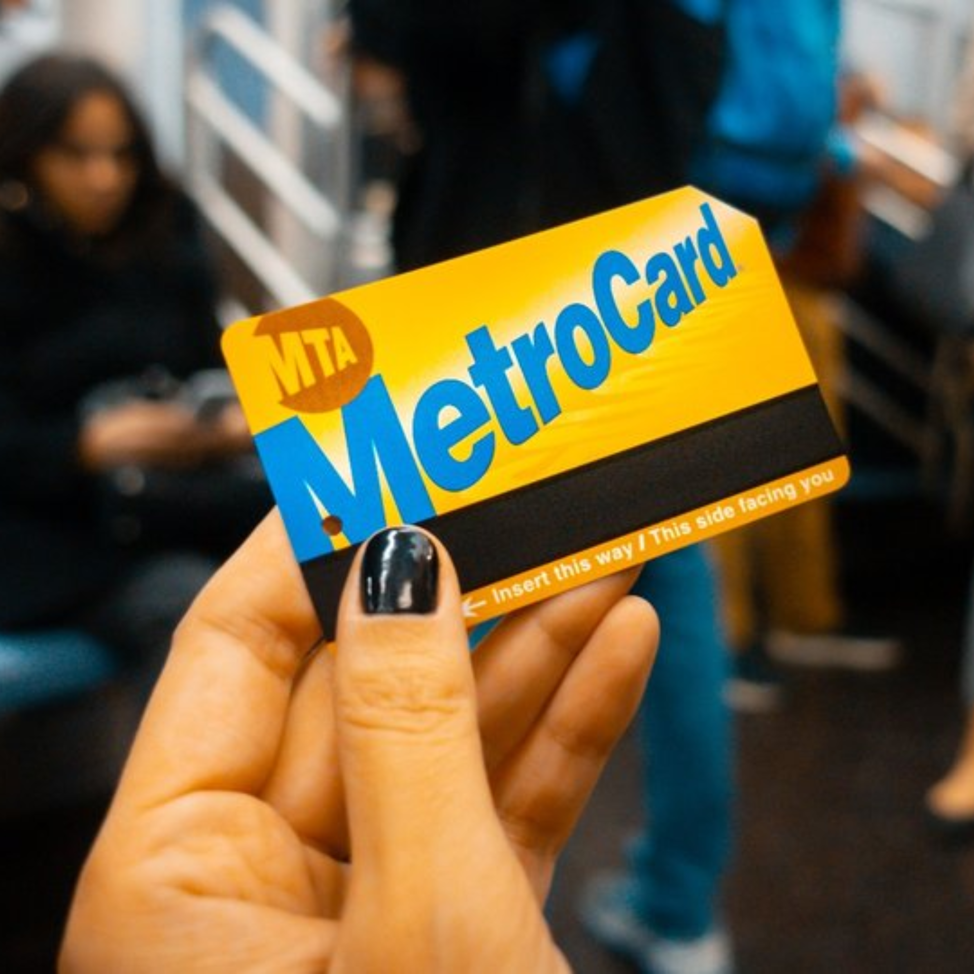 A hand with black nail polish holds up a classic yellow and blue New York City MTA MetroCard inside a subway car. The background is softly blurred, showing other passengers and the train interior.