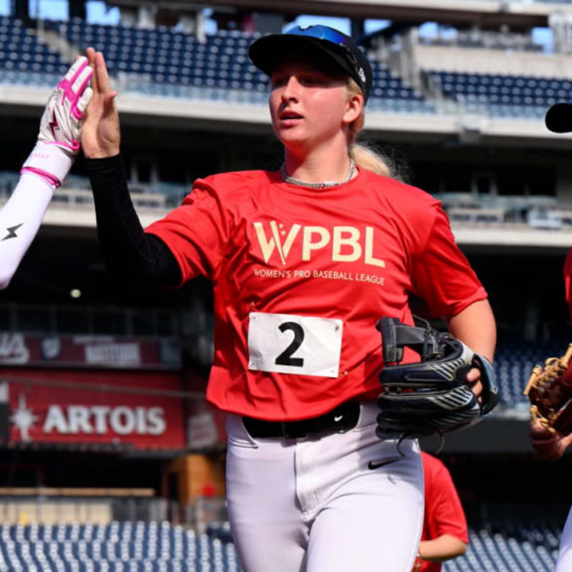 A female baseball player in a red Women’s Pro Baseball League (WPBL) shirt with the number 2 pinned to it gives a high-five to a teammate. She wears white pants, a black cap, and holds a glove while standing on a professional baseball field