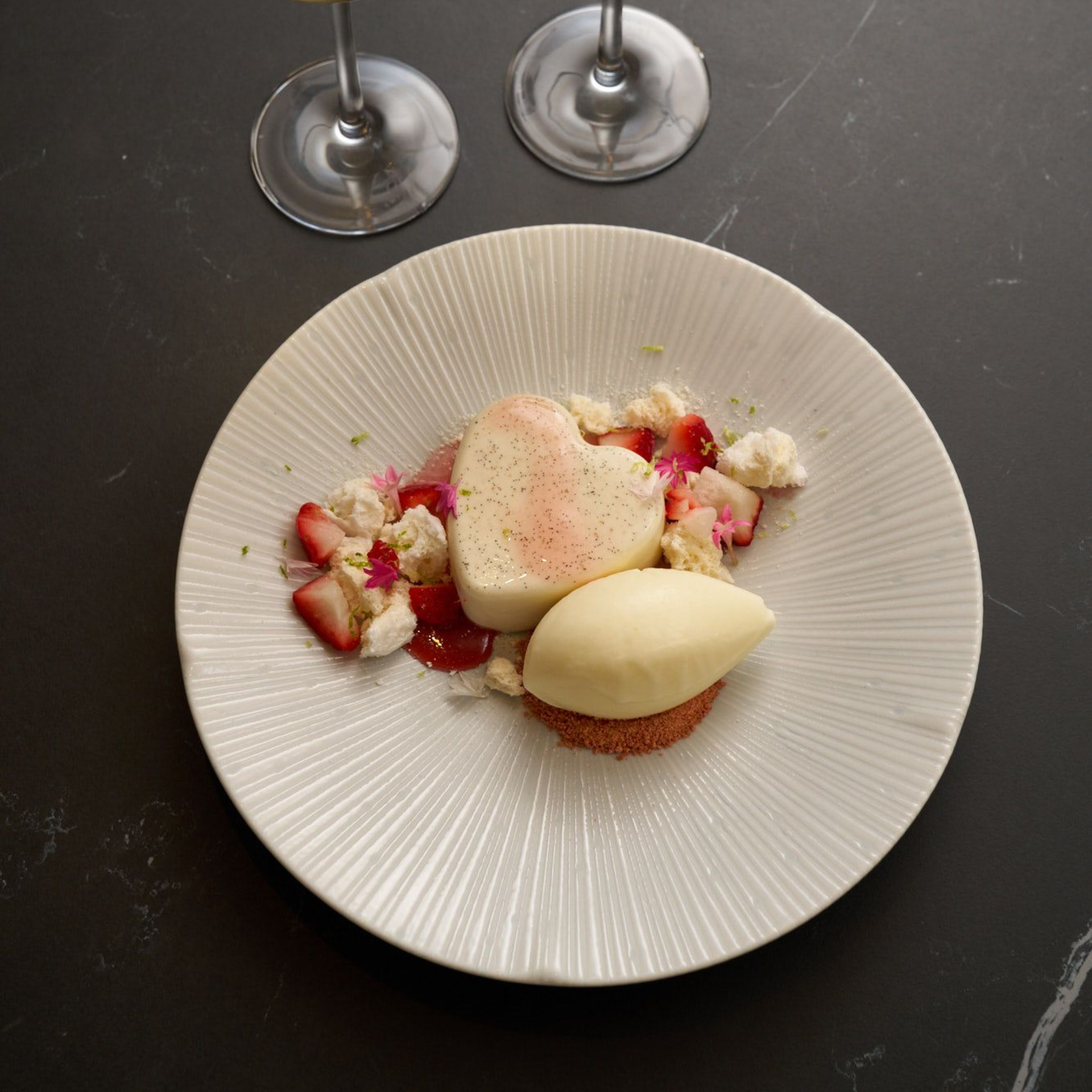 White plate with a pink heart‑shaped dessert, ice cream quenelle on crumbs, strawberries, and small pink flowers