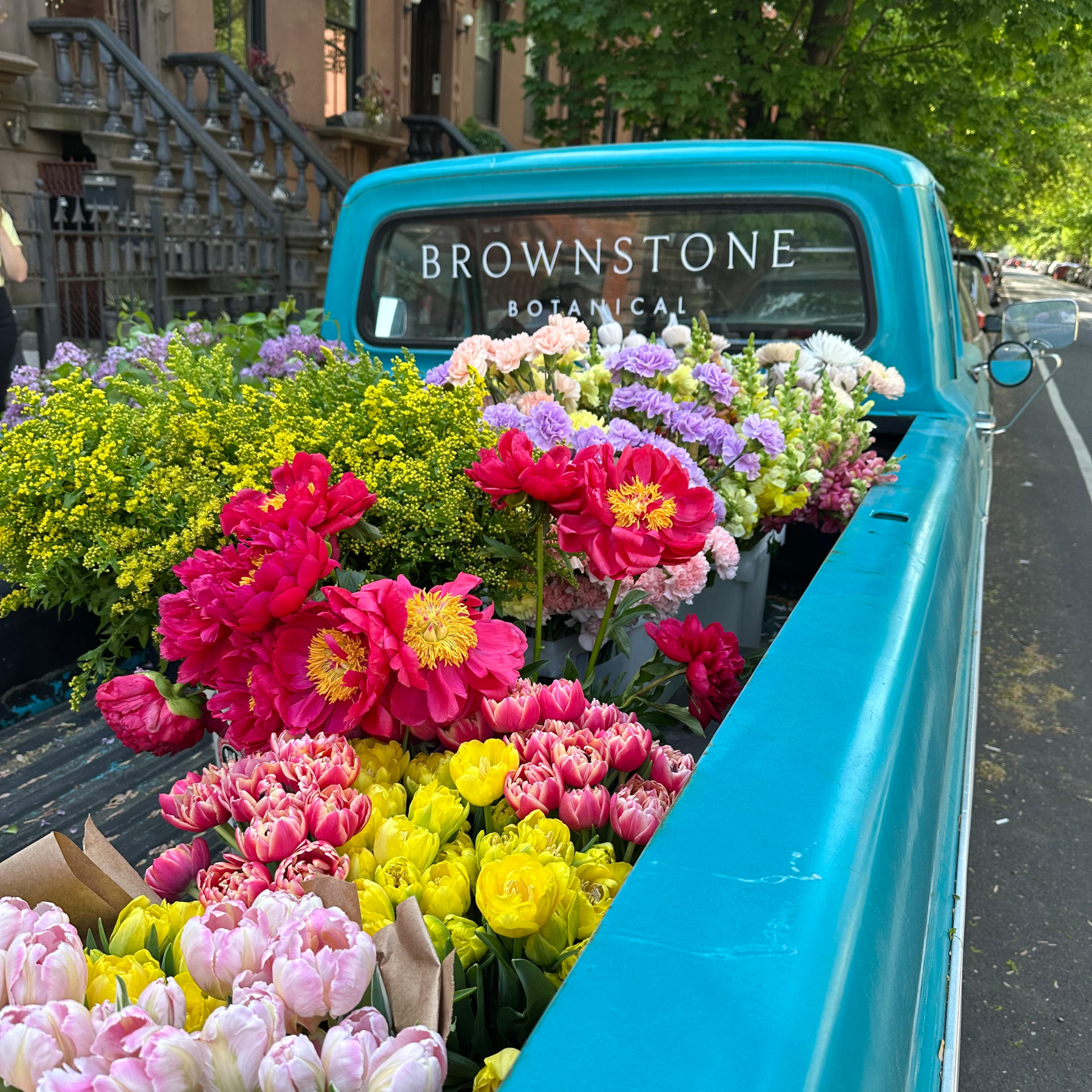 Turquoise pickup truck with bed full of colorful flowers parked on city street near brownstones
