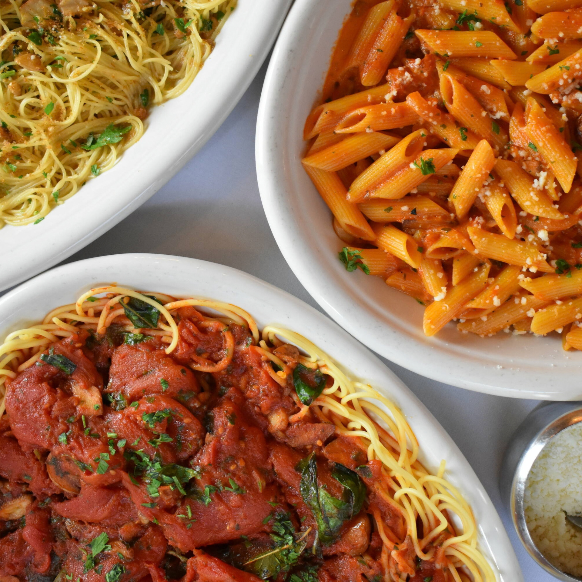 Three oval dishes of pasta—angel hair with garlic and herbs, penne with tomato meat sauce, and spaghetti with basil—served on a white tablecloth with Parmesan and red pepper on the side