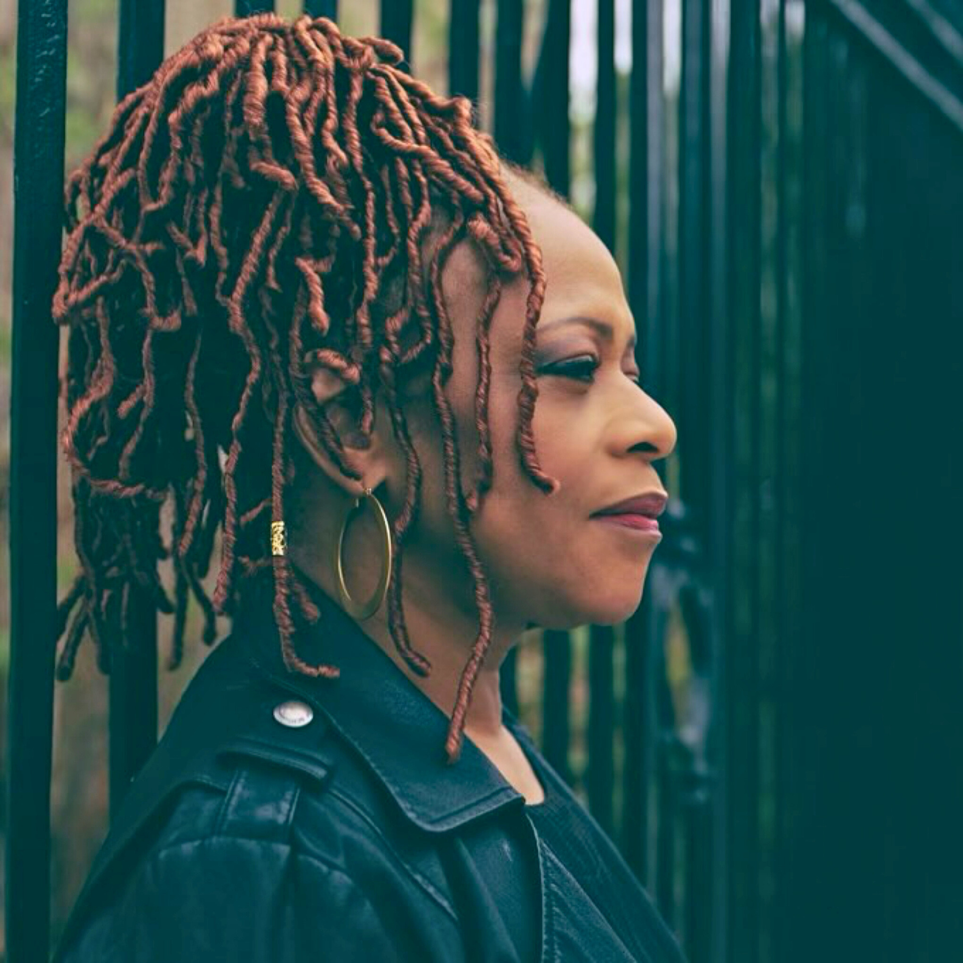 Profile portrait of person with loc-style hair, hoop earrings, and leather jacket near metal fence
