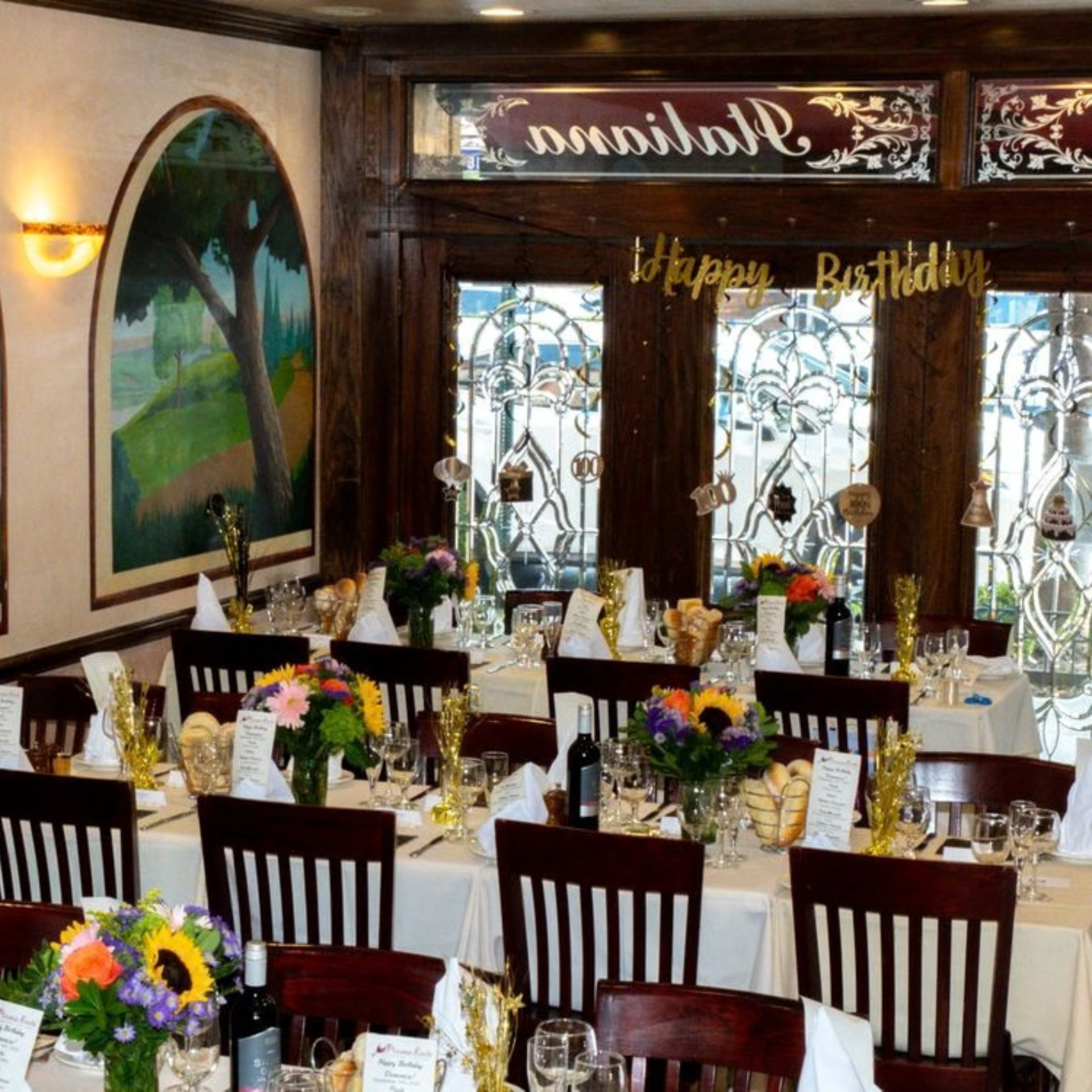 Elegant restaurant dining area decorated for a birthday with white tablecloths, wine bottles, floral centerpieces, and golden “Happy Birthday” signs