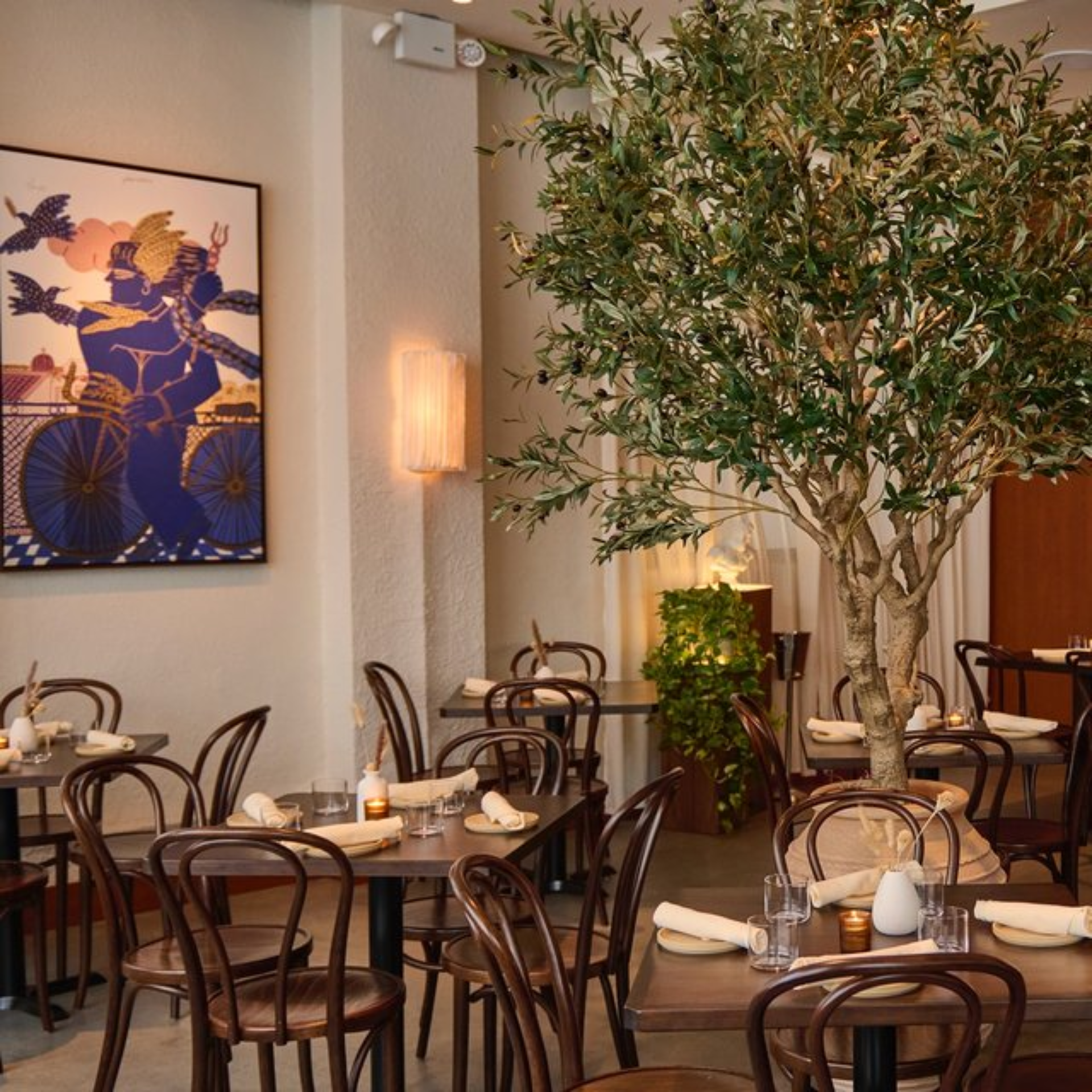 Warm restaurant interior with wooden tables, indoor tree, and framed artwork on the wall