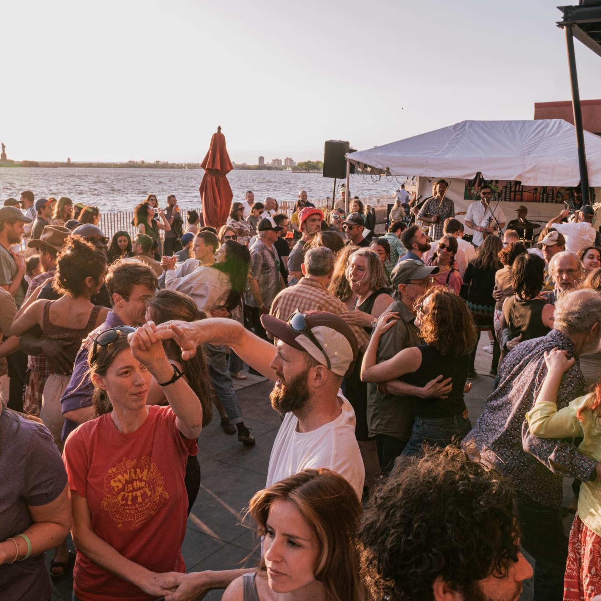 Outdoor waterfront gathering at sunset with people dancing, musicians under canopy, and city skyline view