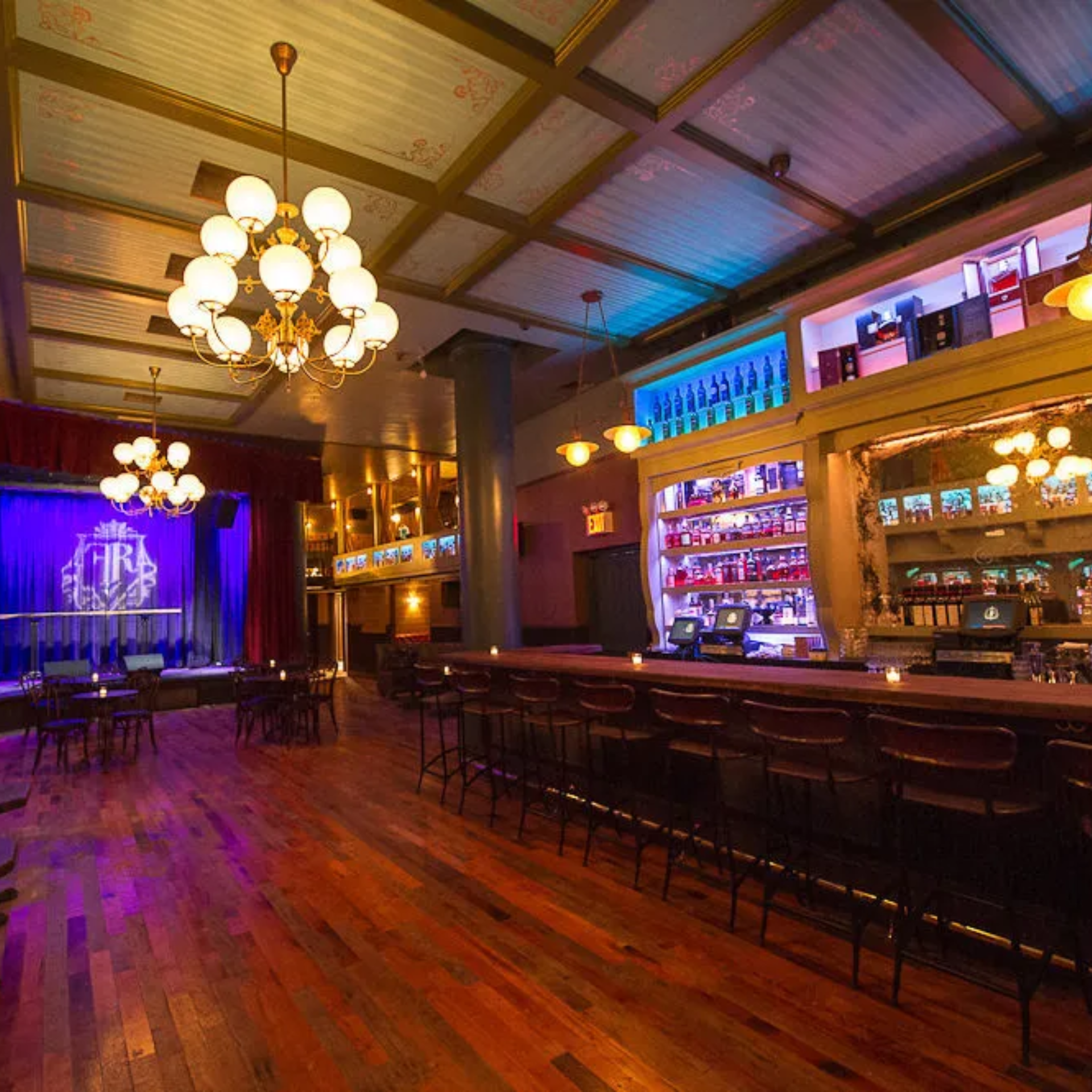 Stylish bar with chandeliers, wooden counter, stocked shelves, and stage with blue curtain
