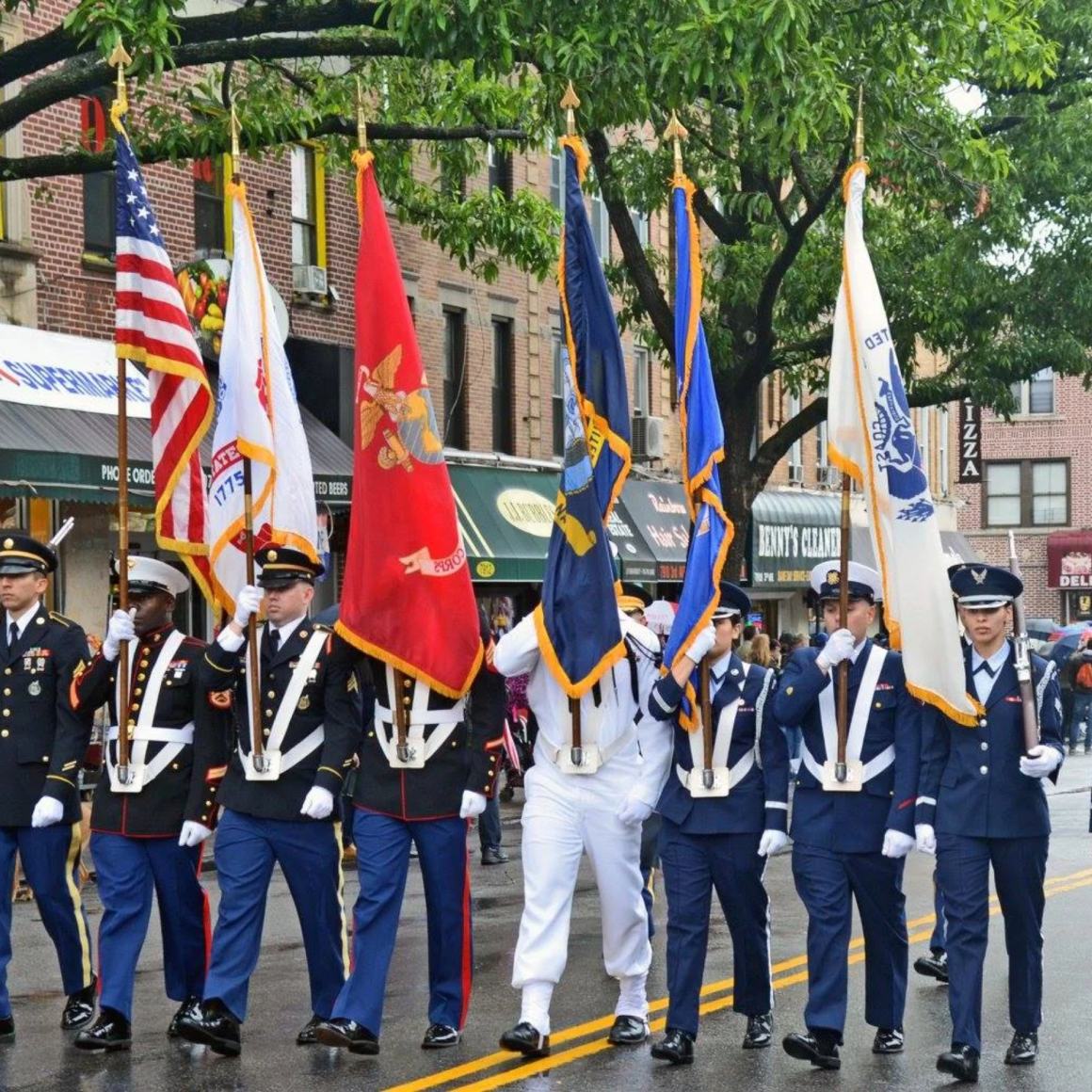 Military parade in city street with U.S. Armed Forces flags and uniformed personnel marching past storefronts after rain