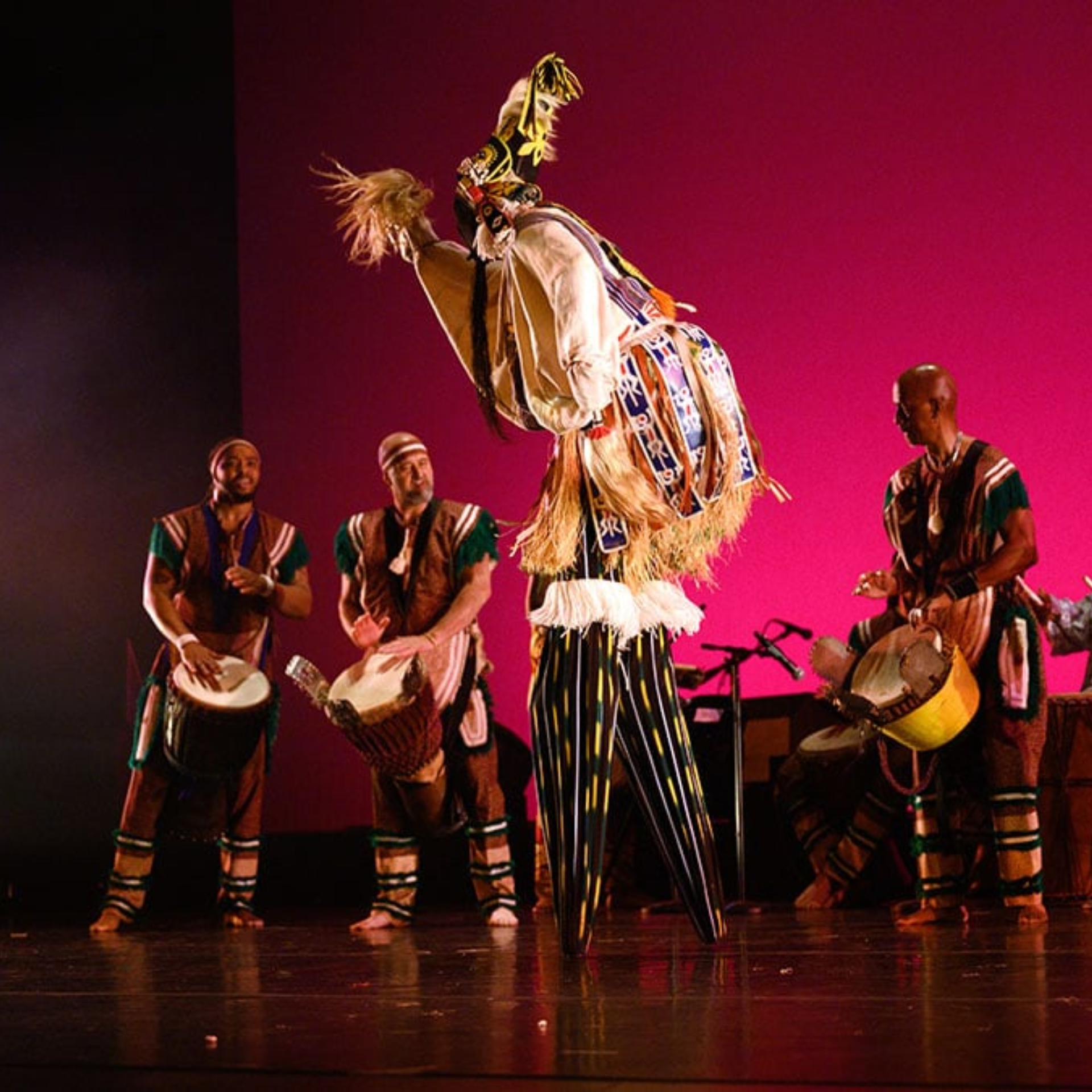Traditional African cultural dance performance with masked dancer in colorful costume and drummers on stage under vibrant pink lighting