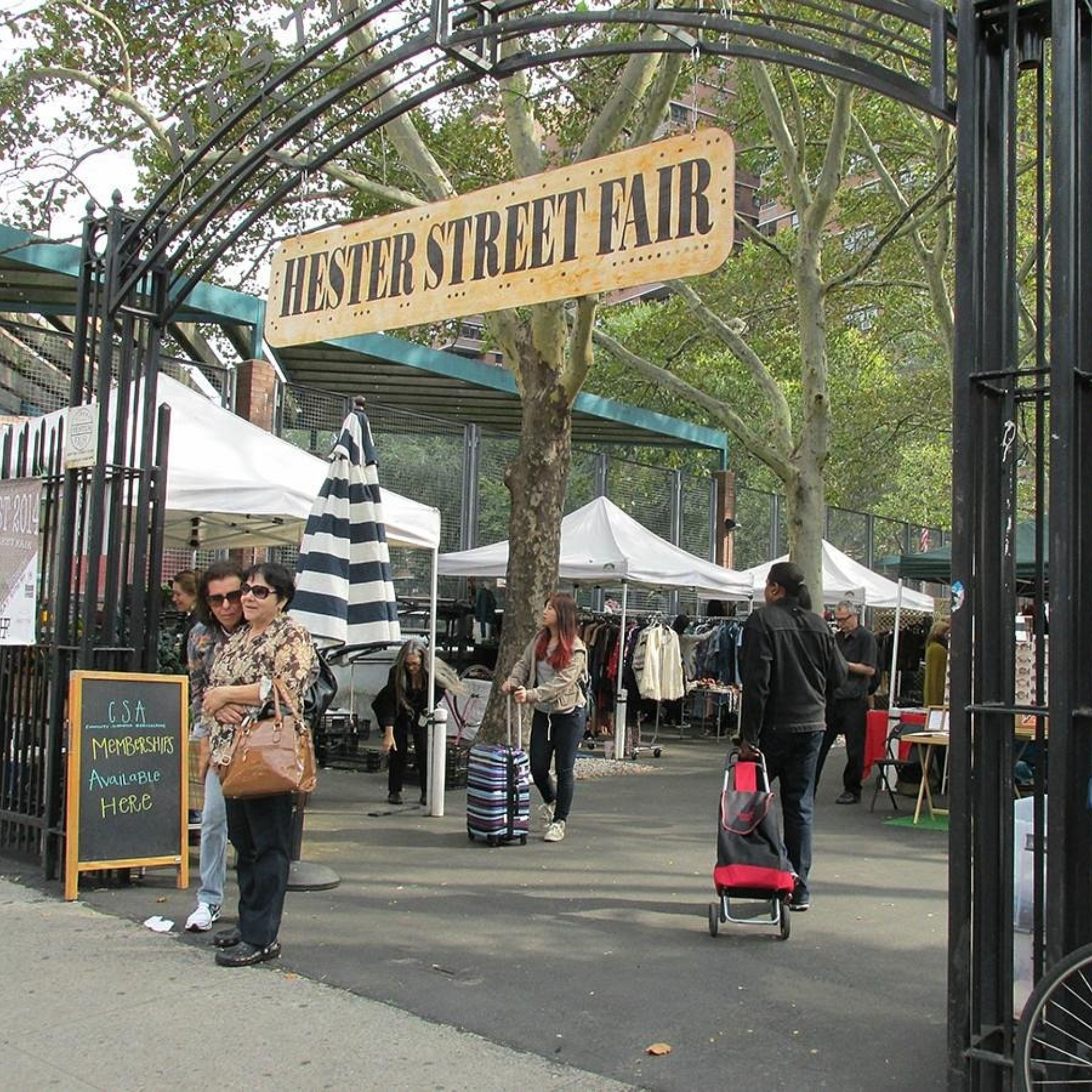 Hester Street Fair entrance archway with wooden sign, vendor tents, and visitors shopping at outdoor NYC community market