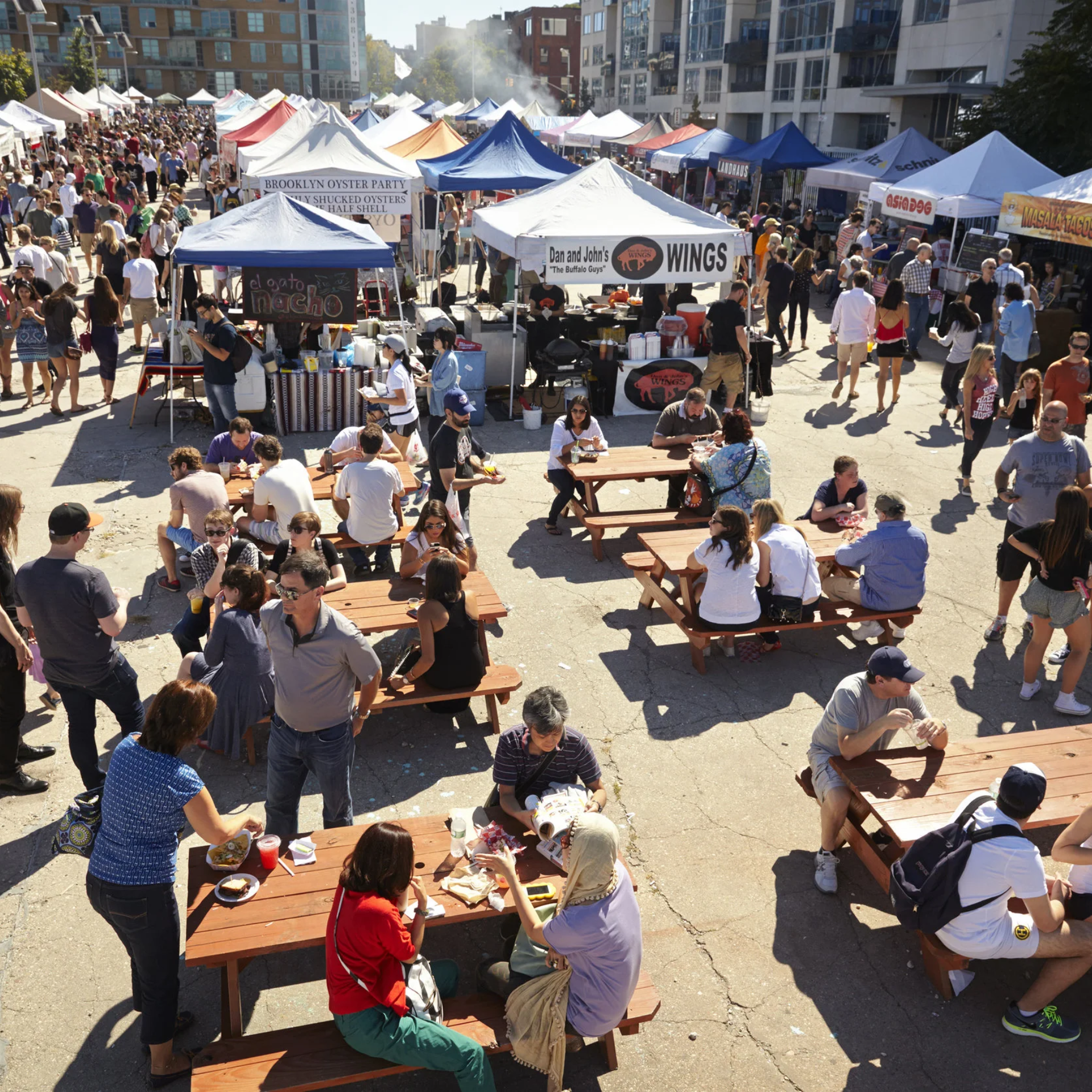 Outdoor NYC food festival with vendor tents, diverse cuisine stalls, and crowds dining at picnic tables in lively urban setting