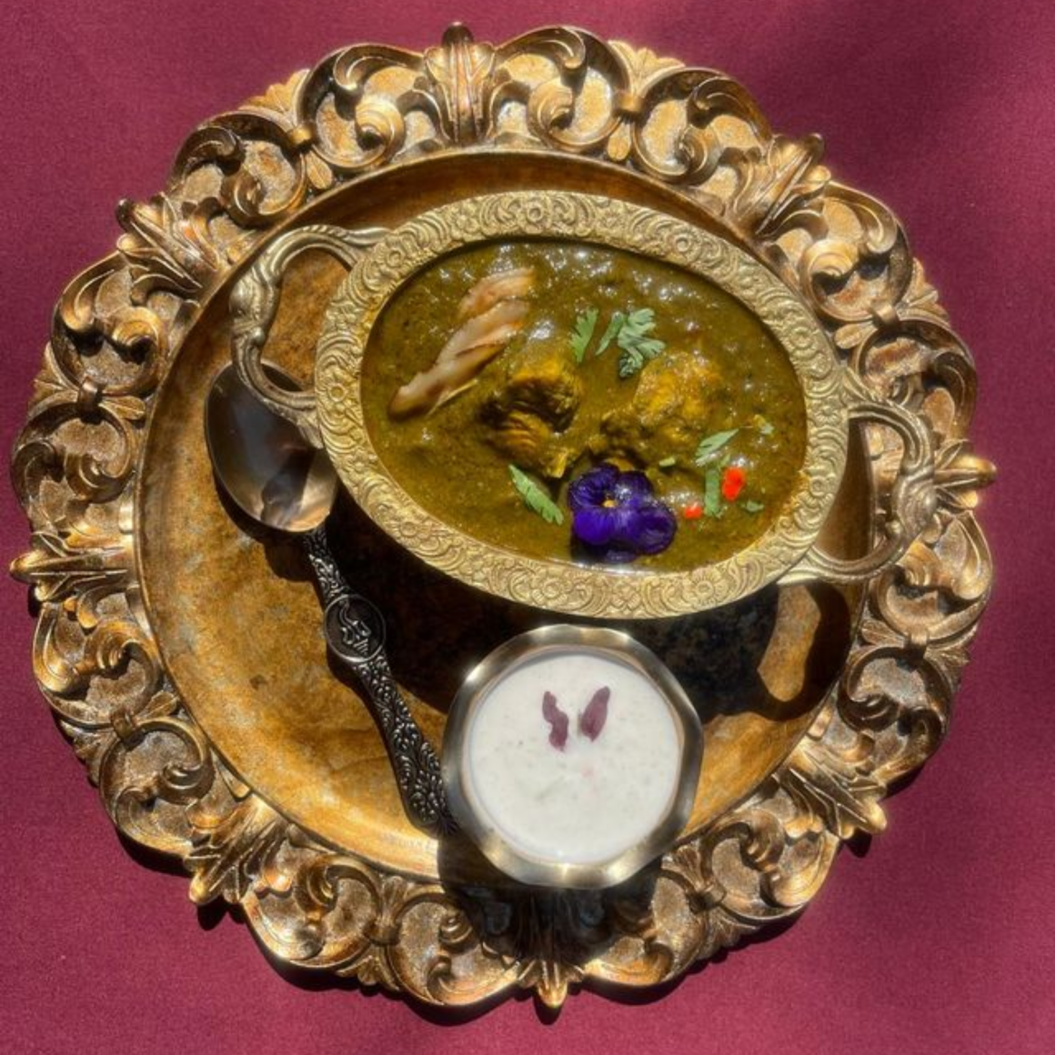 Elegant golden plate with ornate bowl of green curry, yogurt sauce, and silver spoon on maroon surface