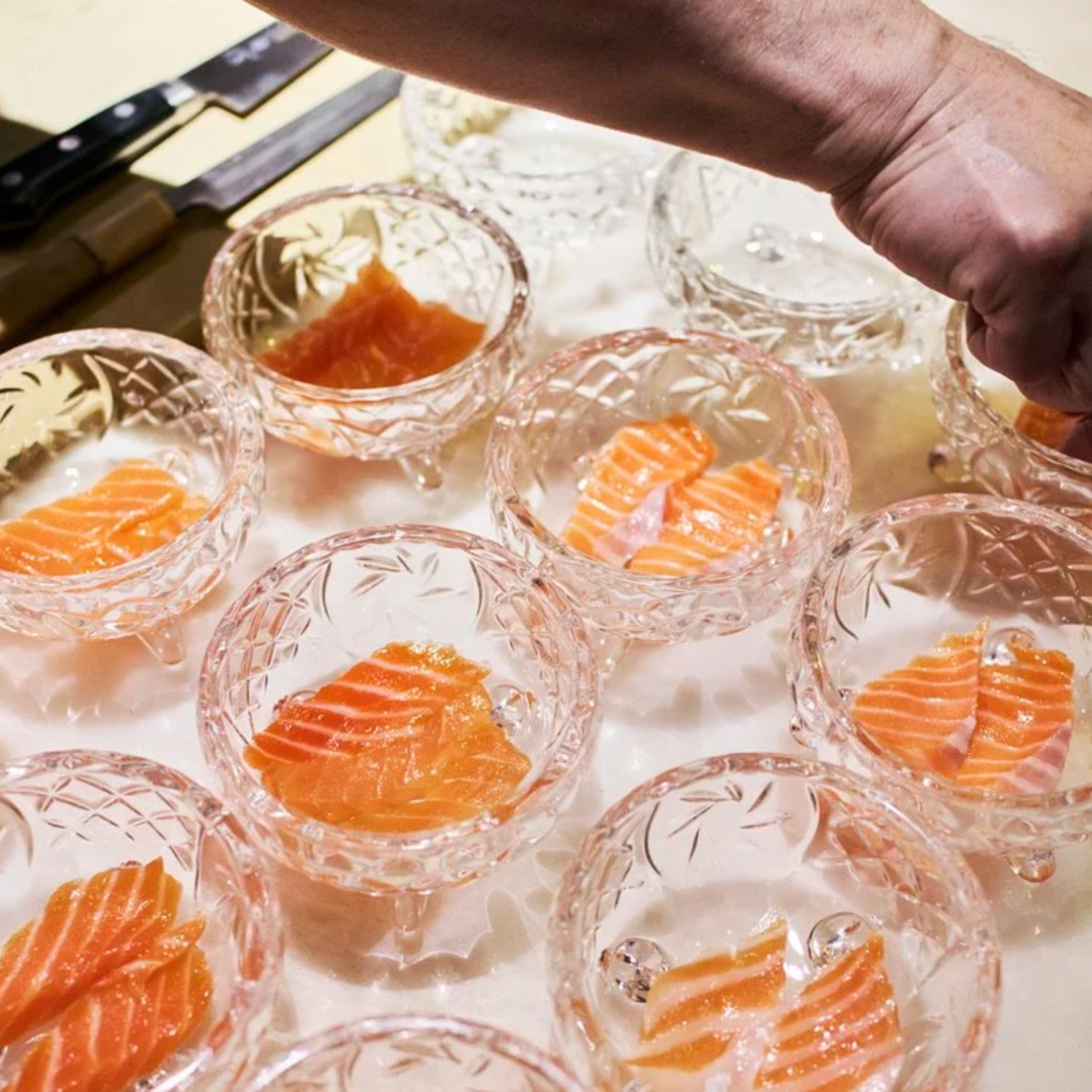 Glass bowls of salmon sashimi being plated by hand in kitchen setting