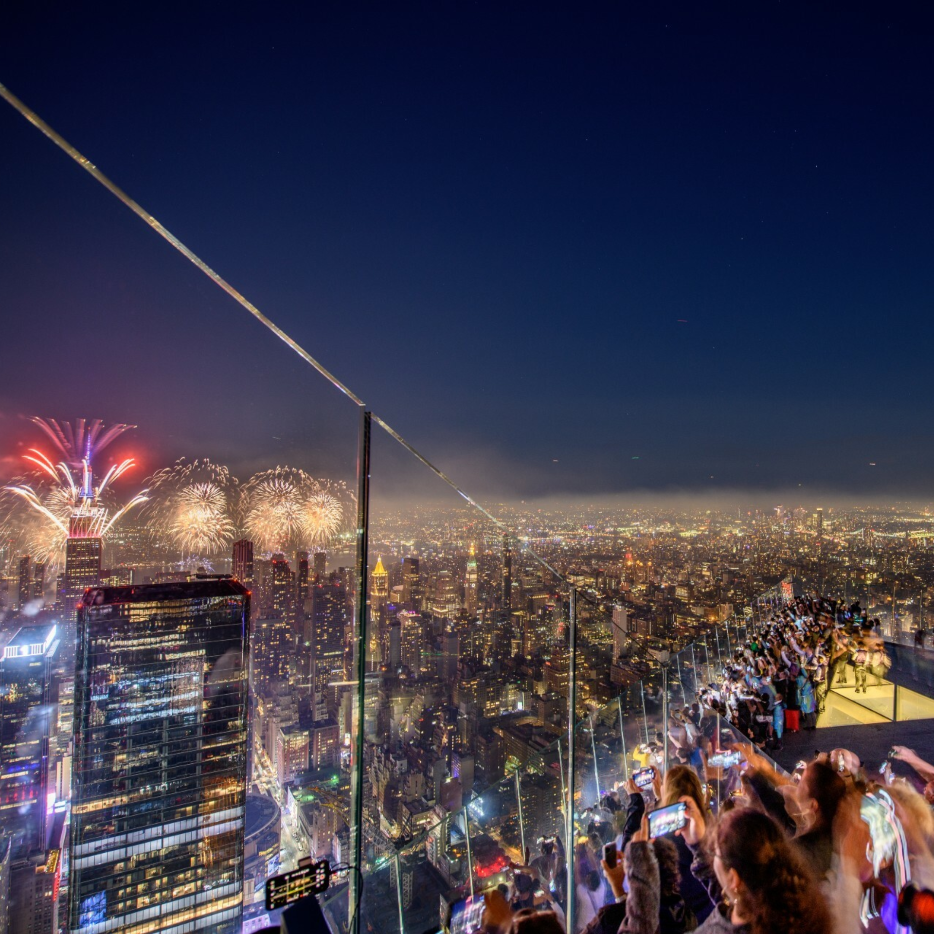 Nighttime city view from high observation deck with crowd behind glass barriers, people filming illuminated skyline and red, white, gold fireworks bursting overhead