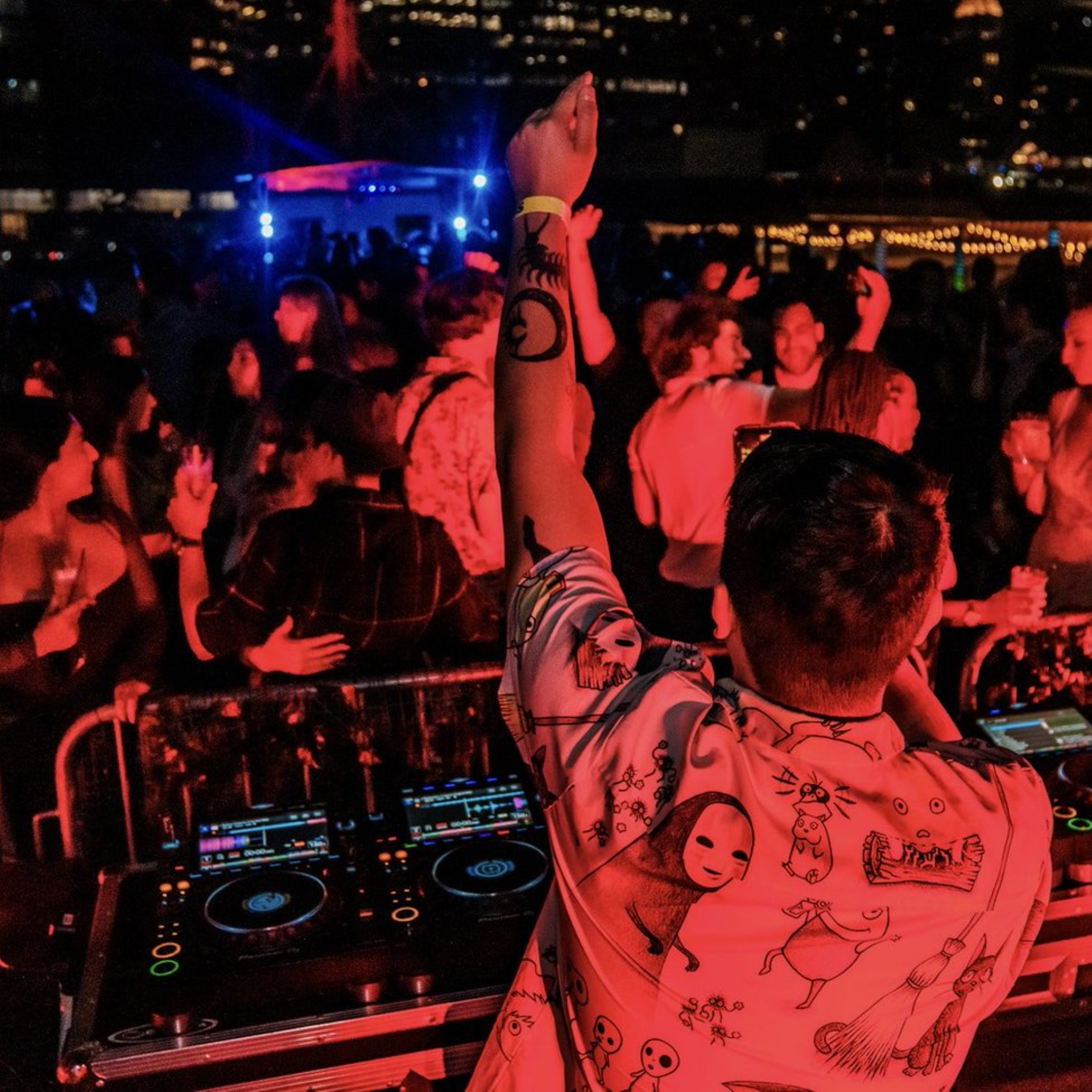 DJ raising arm at urban rooftop party, mixing console with turntables, crowd dancing under red and blue lights, city skyline backdrop