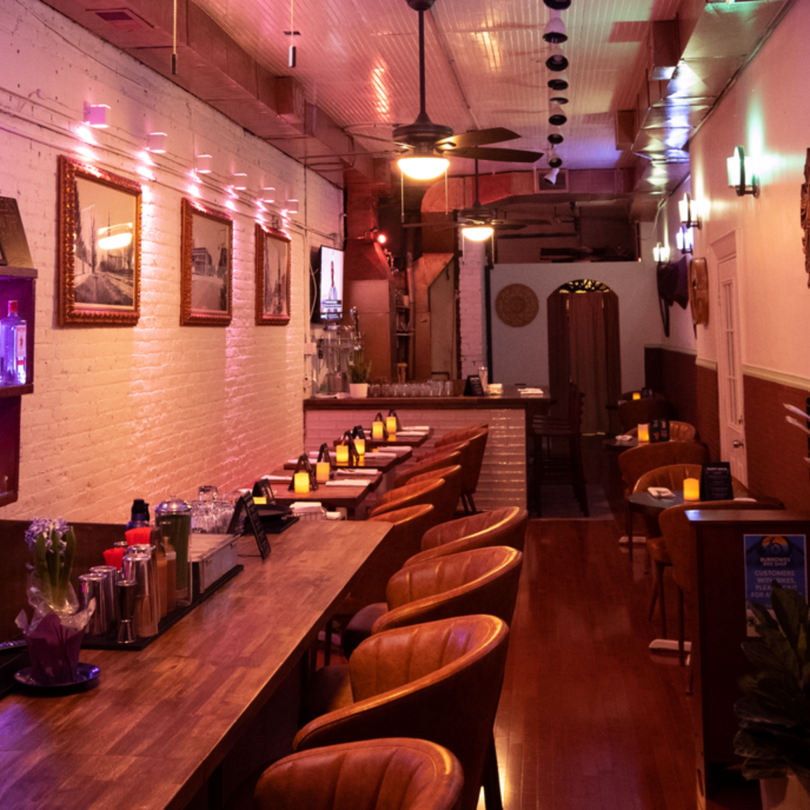 Cozy bar interior with wooden counter, leather chairs, warm ambient lighting, and shelves of bottles against white brick walls