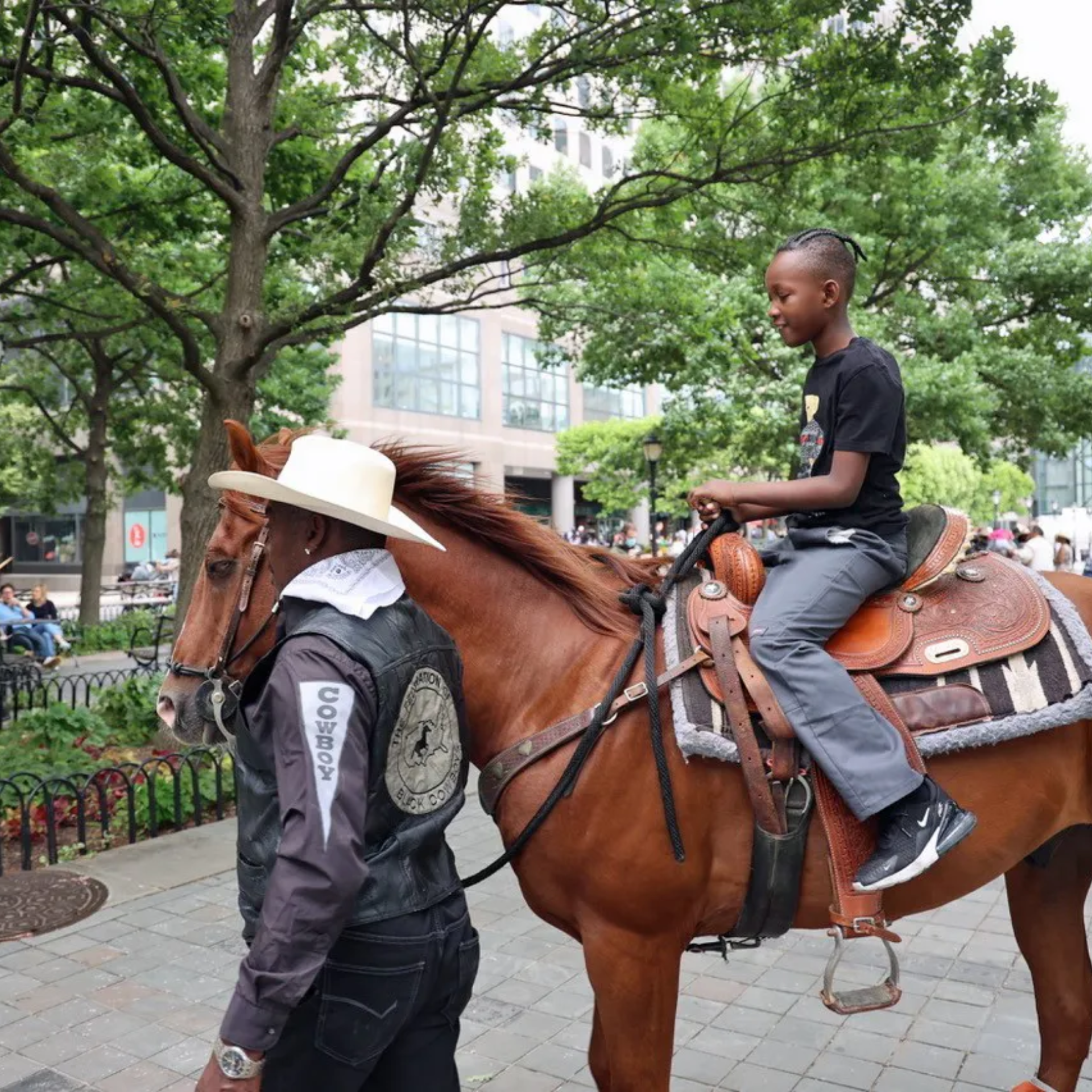 Child riding brown horse in urban park, cowboy guide in hat and vest leading