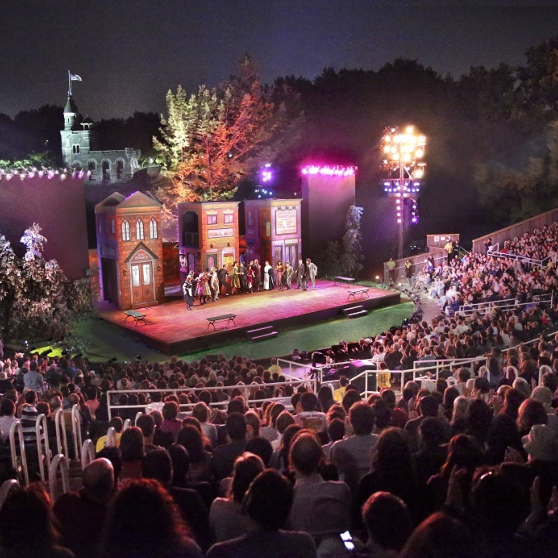 Open‑air night theater performance with colorful stage lights, town street set, large cast, and audience in amphitheater under scenic backdrop