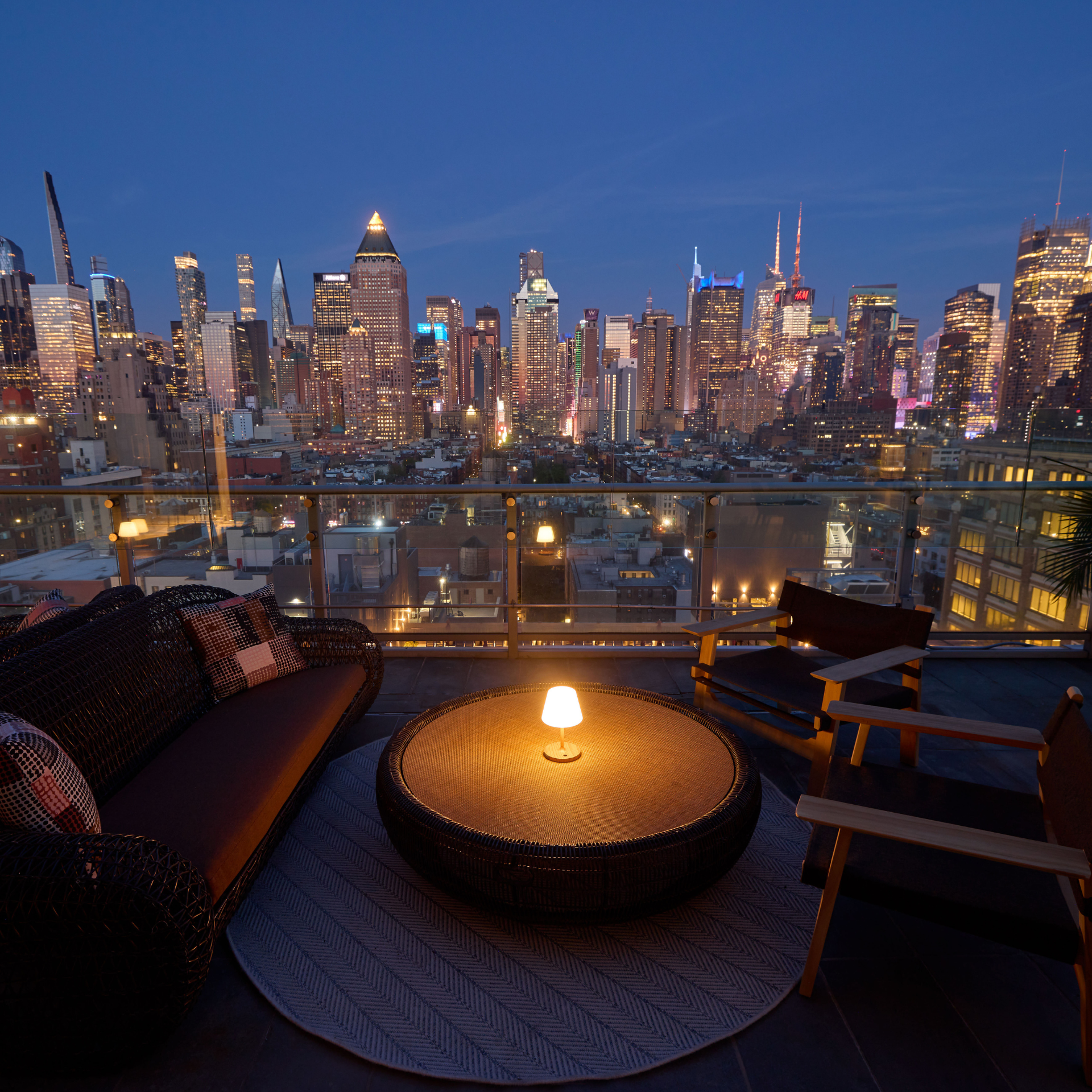 Rooftop terrace at dusk with woven sofa, patterned cushions, wooden chairs, glowing lamp on round table, and panoramic city skyline illuminated by building lights