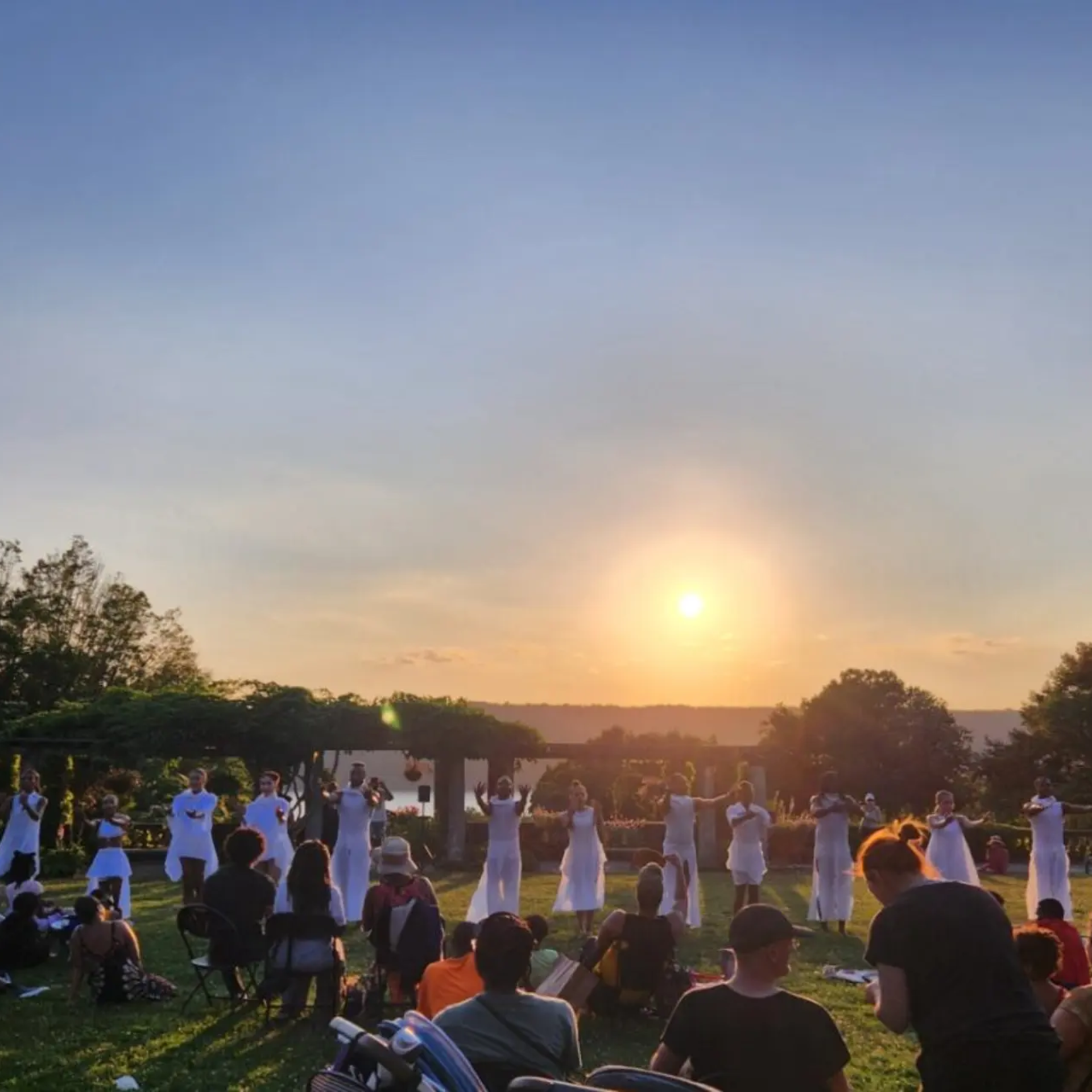A group of dancers dressed in white performing outdoors on a grassy lawn at sunset, with an audience watching from the grass in the foreground