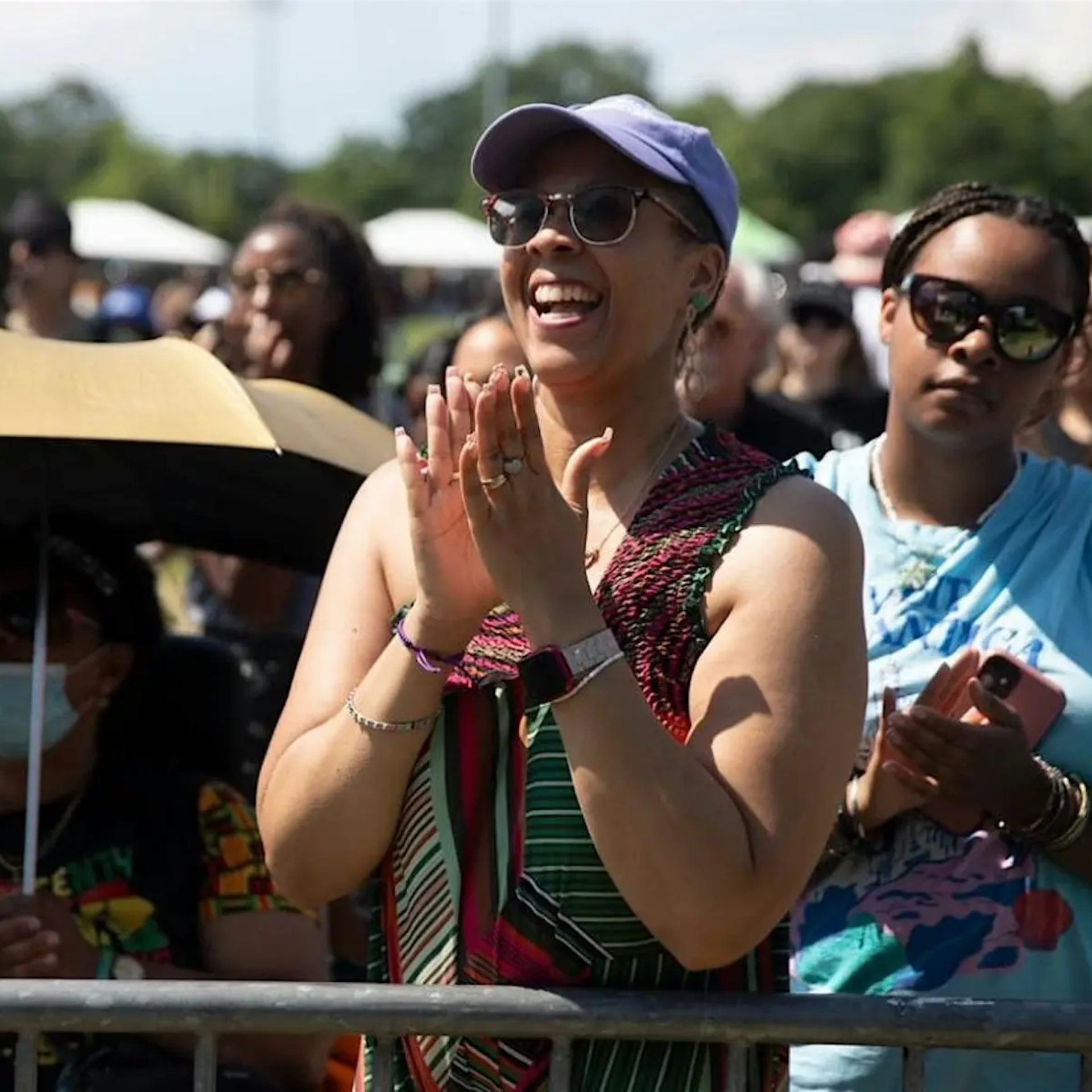 Festival crowd clapping outdoors, person in purple cap and colorful patterned outfit celebrating under sunny tents