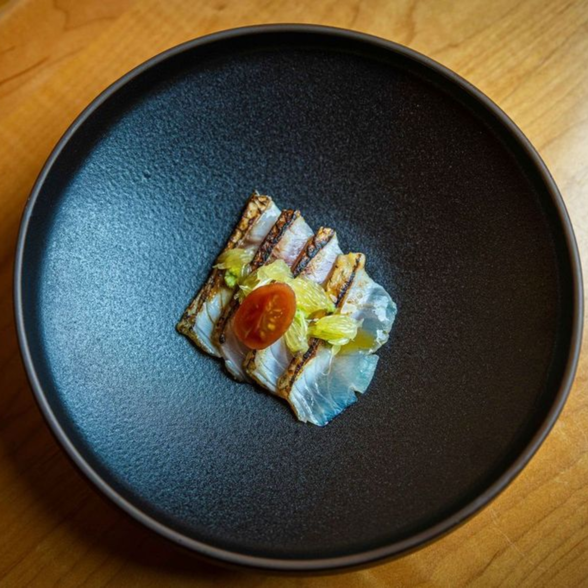 Minimalist fine dining plate with seared fish slices, tomato garnish, and green vegetables on dark textured dish