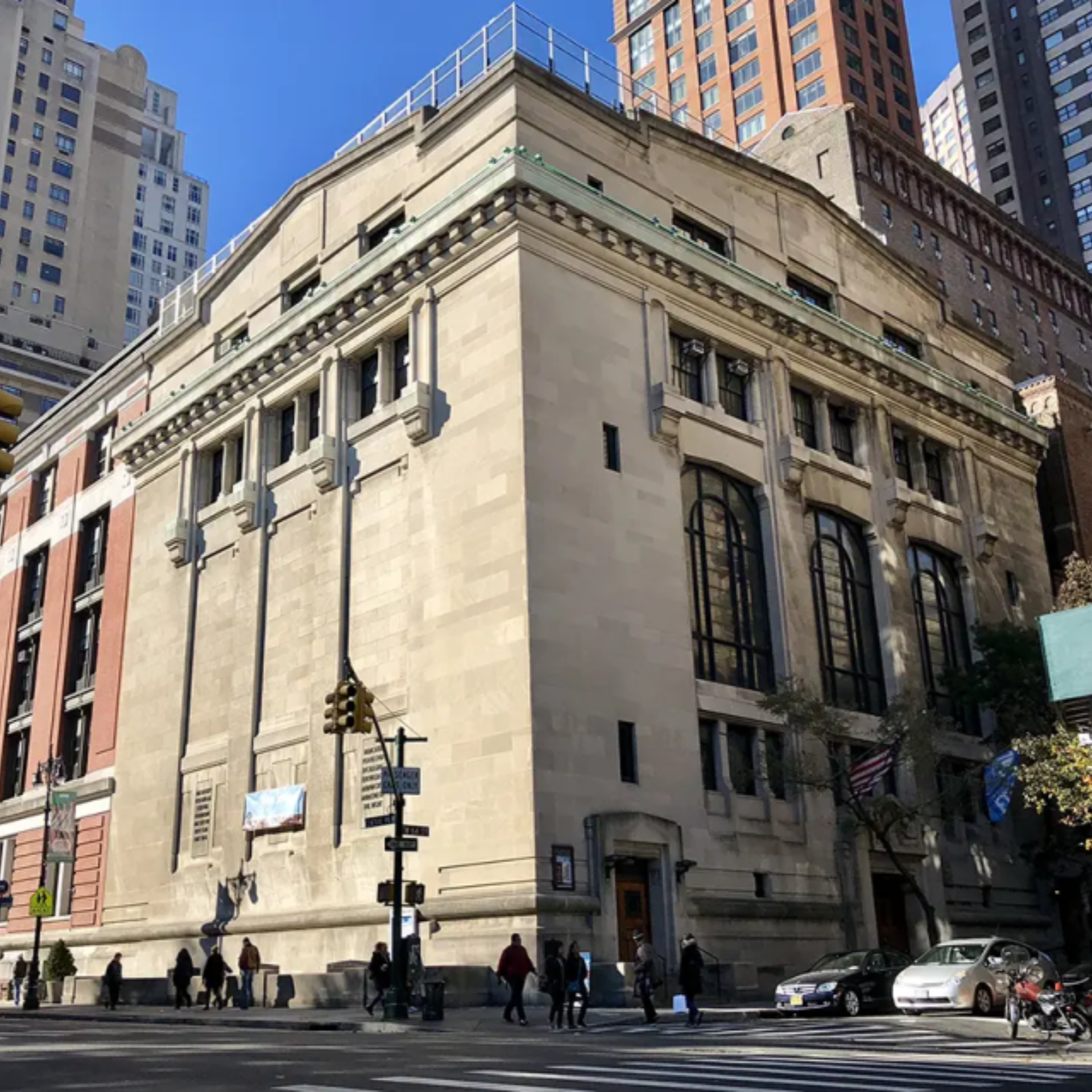 Historic stone building with arched windows on city street corner, surrounded by modern high‑rises under blue sky