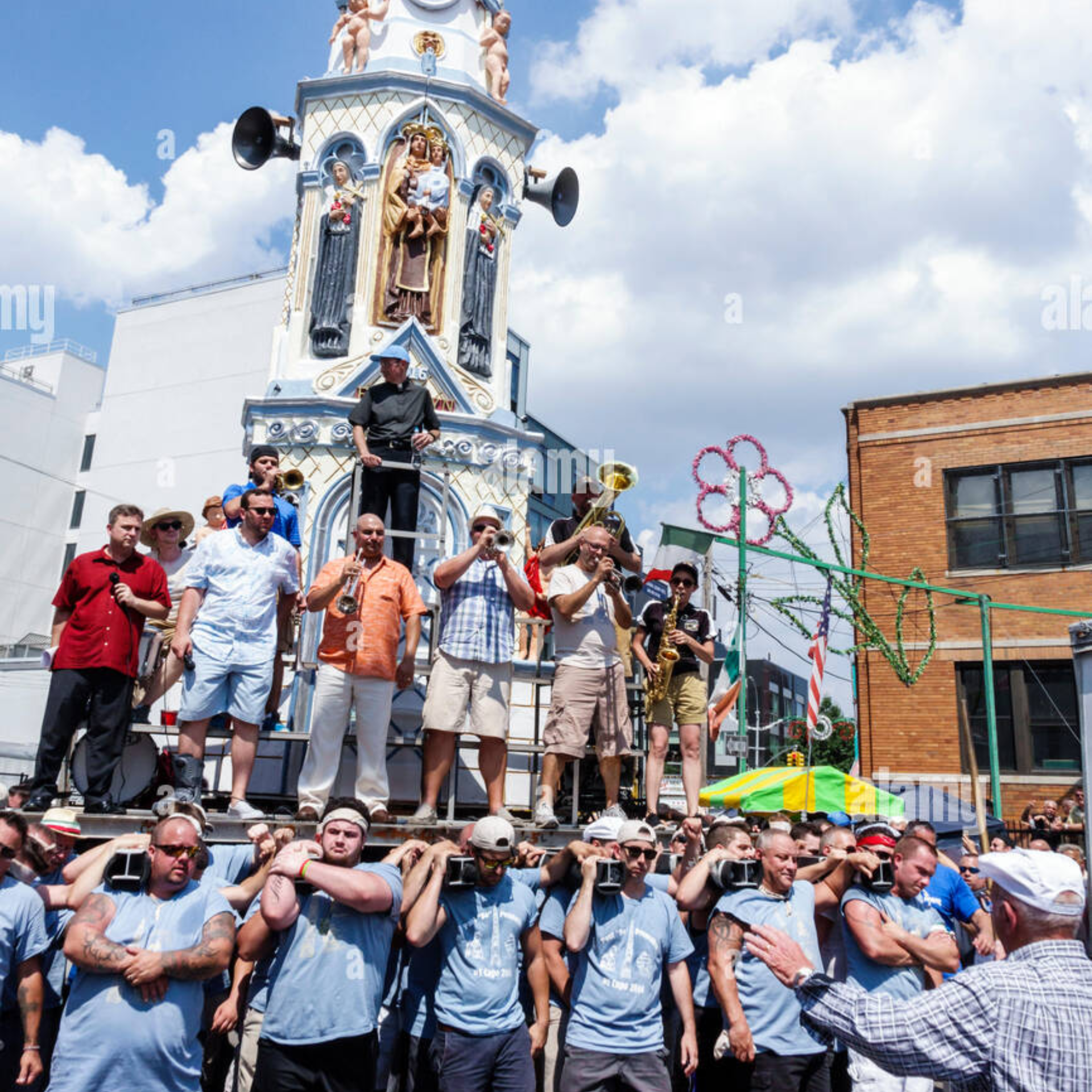 Religious street procession with ornate Virgin Mary statue, musicians playing brass instruments, and men in blue shirts carrying platform under bright sky