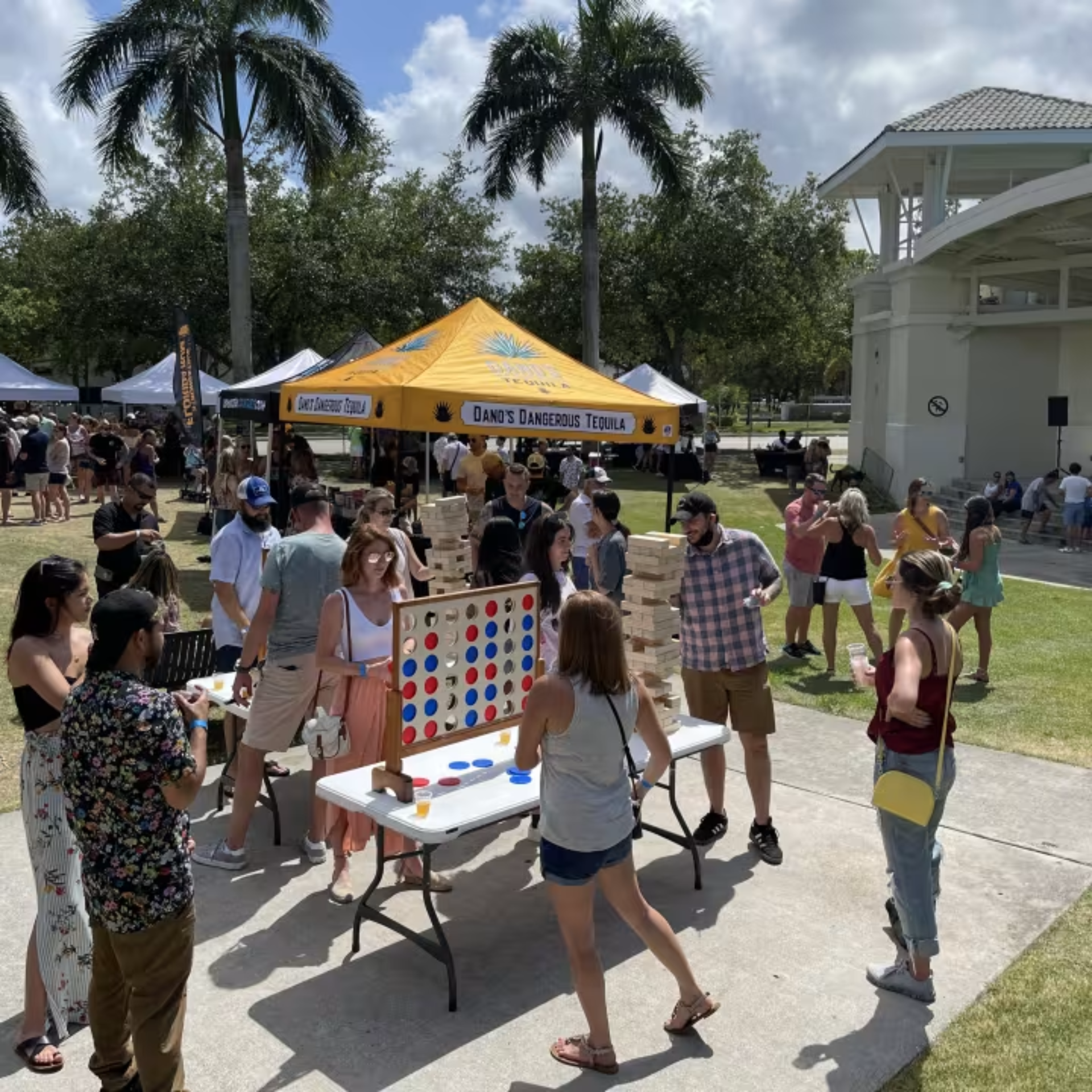 Outdoor festival in park with palm trees, giant Connect Four and Jenga games, tequila tent, and people enjoying sunny social event