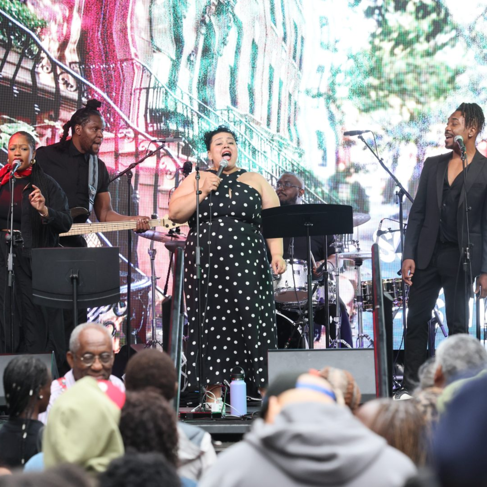 Outdoor live concert with three vocalists and band, singer in polka dot dress performing passionately before crowd