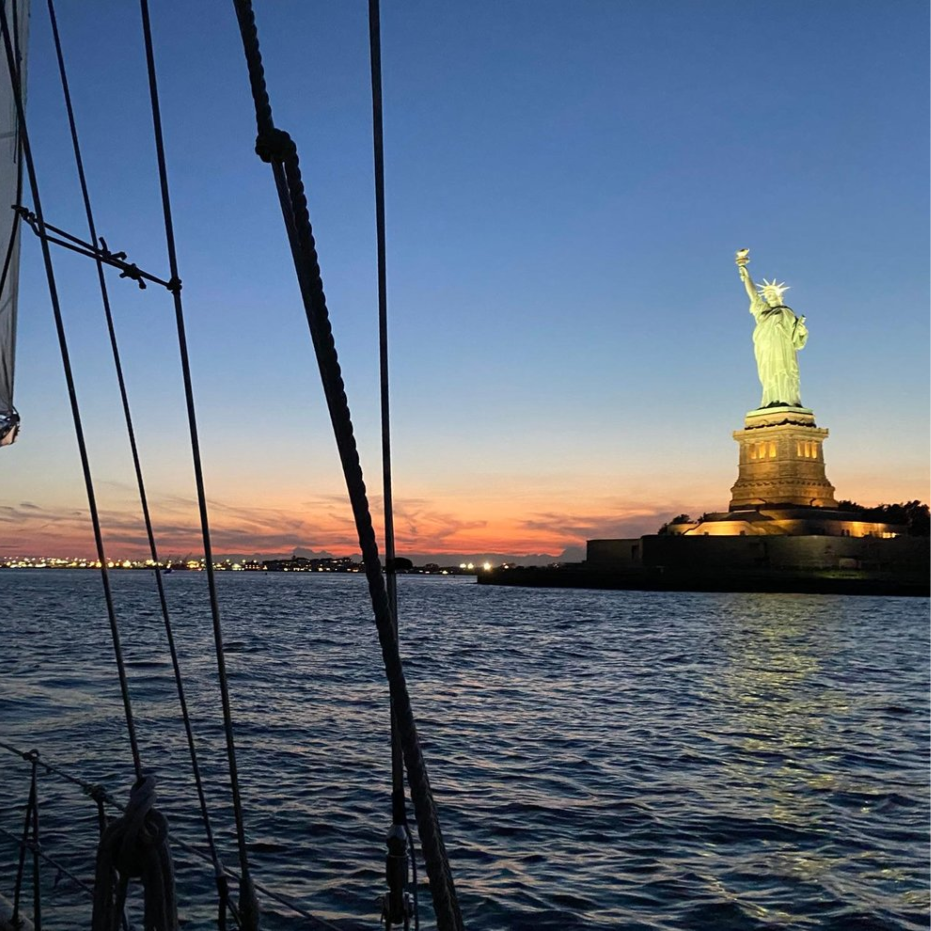 Statue of Liberty glowing at sunset, viewed from boat with rigging and ropes, city lights in background