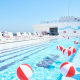 Rooftop swimming pool at JFK with red‑and‑white beach balls, striped lounge chairs, and sunny blue sky