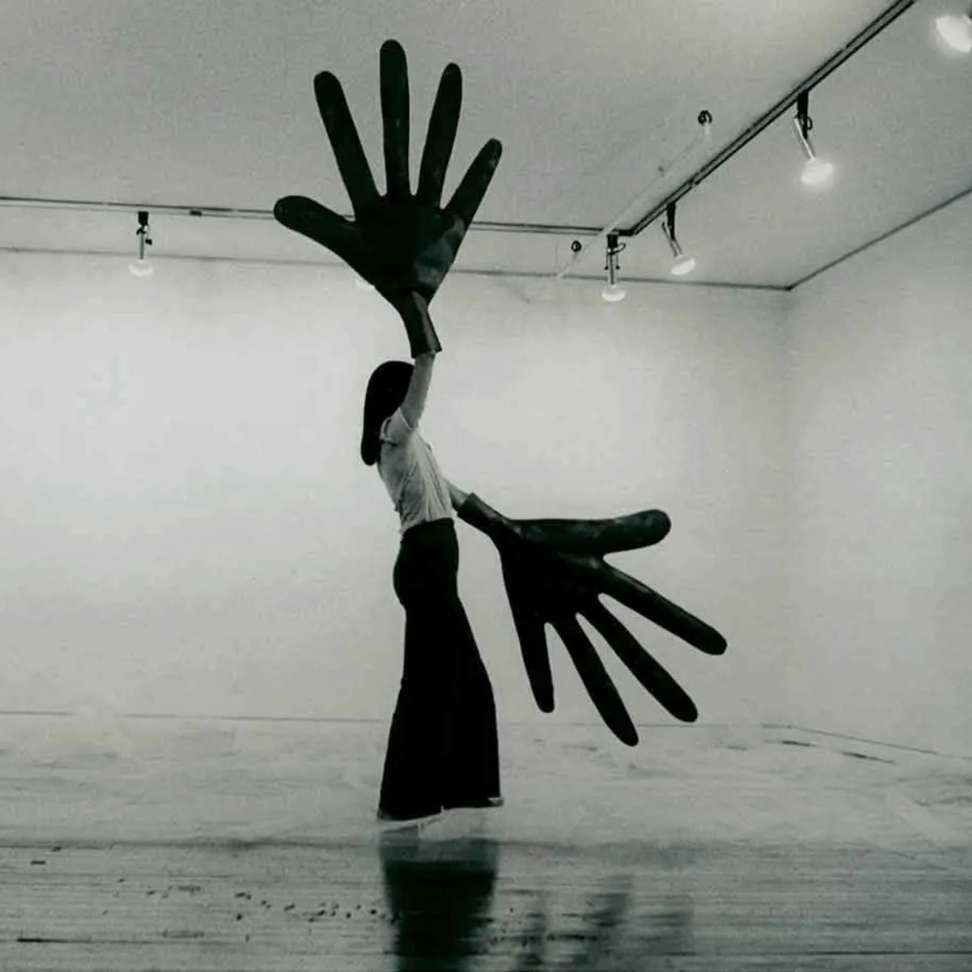 Black-and-white photo of a person holding oversized hand-shaped props in an empty gallery space.