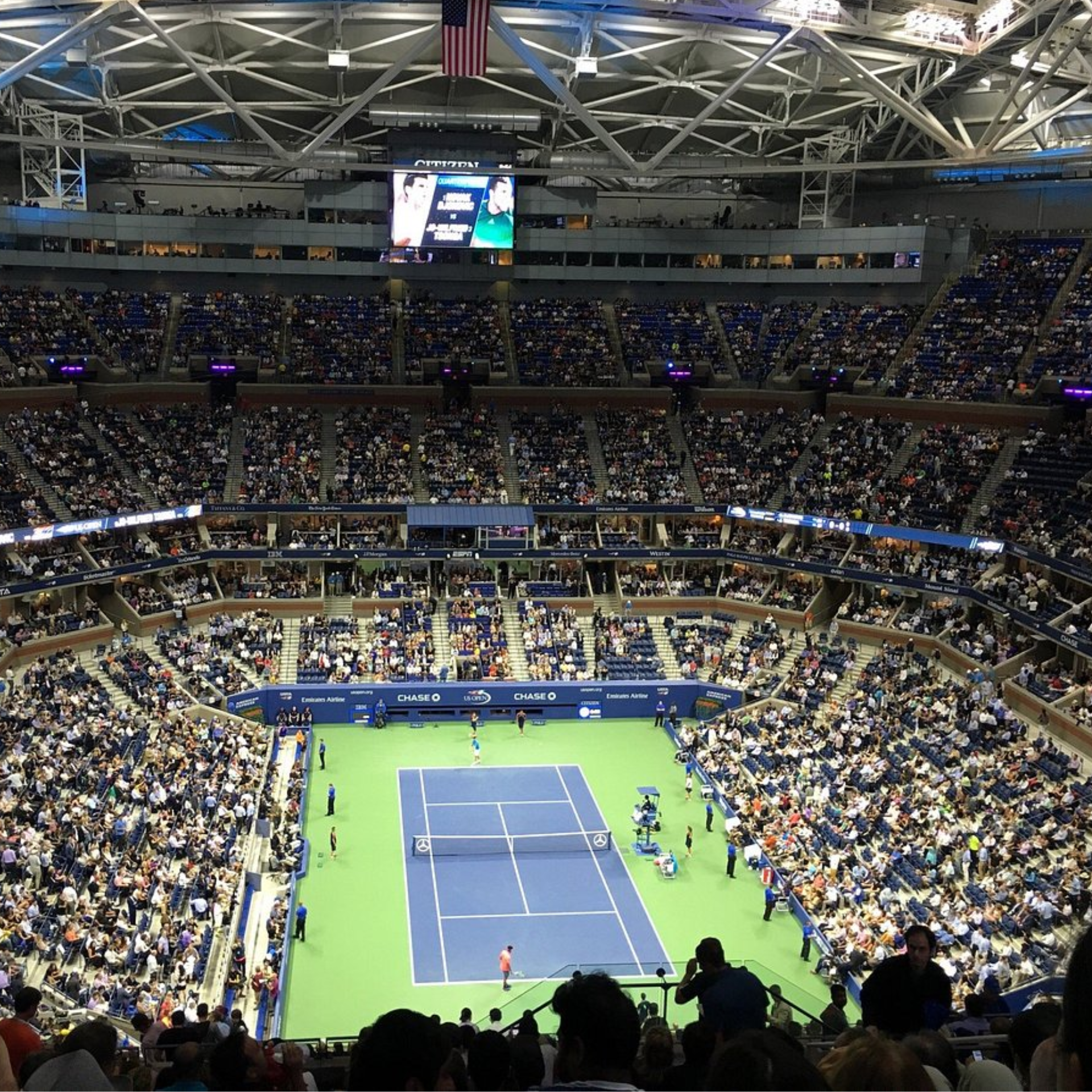 Large NYC tennis stadium with blue court, players competing, electronic scoreboard, and crowd of spectators, major sports event featured in daily things to do in NYC any day