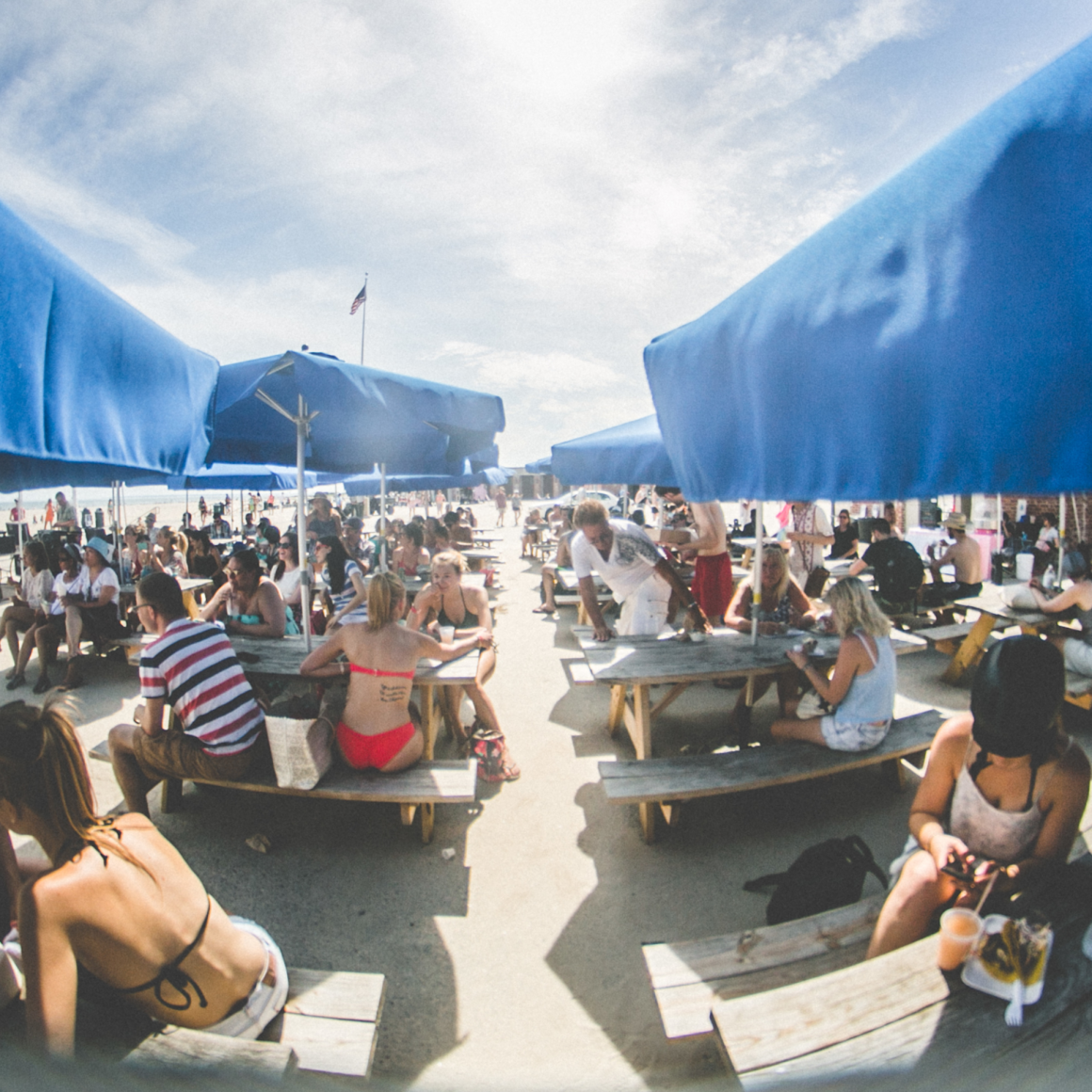 Beachside NYC café with picnic tables, blue umbrellas, and lively crowd in swimsuits on sunny day, coastal leisure spot featured in daily things to do in NYC any day