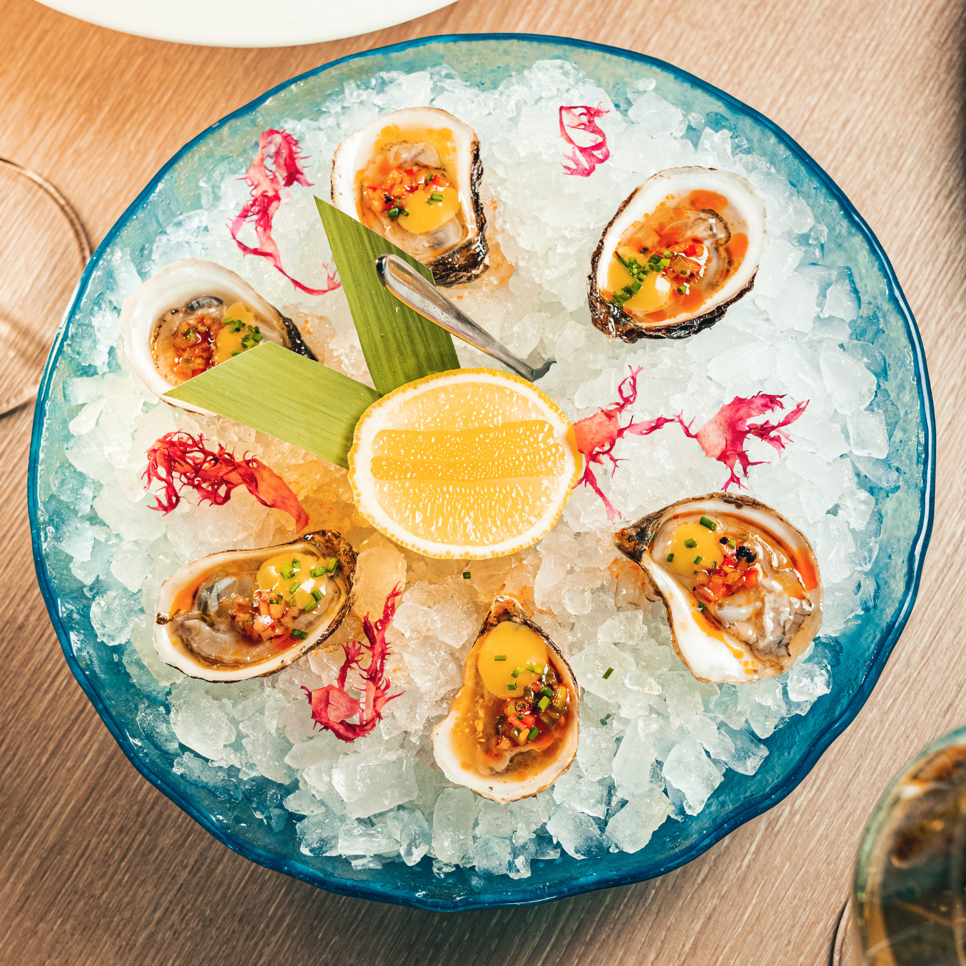 Eight fresh oysters on crushed ice, garnished with yellow-orange sauce, herbs, and red accents lemon and fork at center