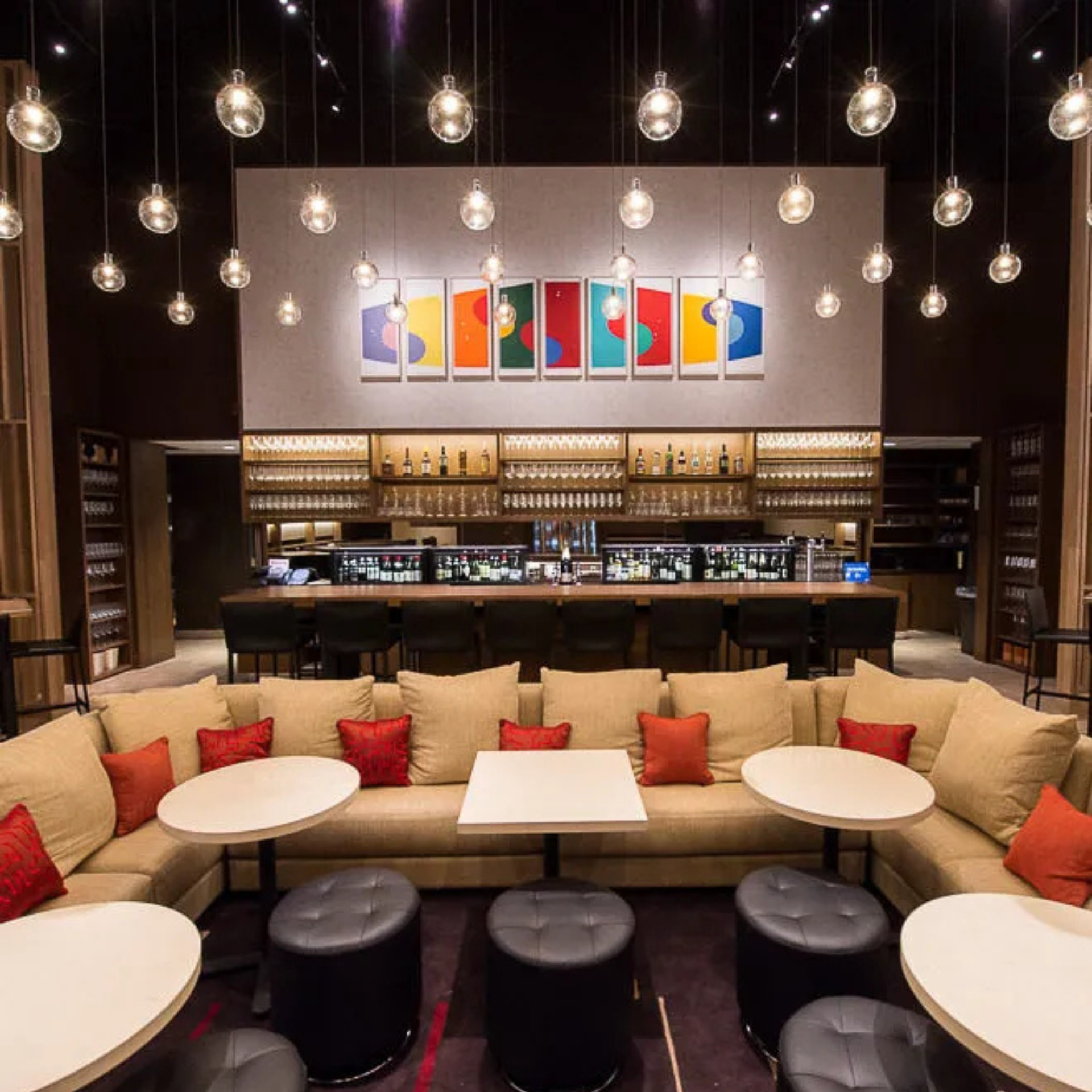 Modern lounge with beige sofas, red pillows, round tables, pendant lights, and vibrant art above a sleek, well-stocked bar