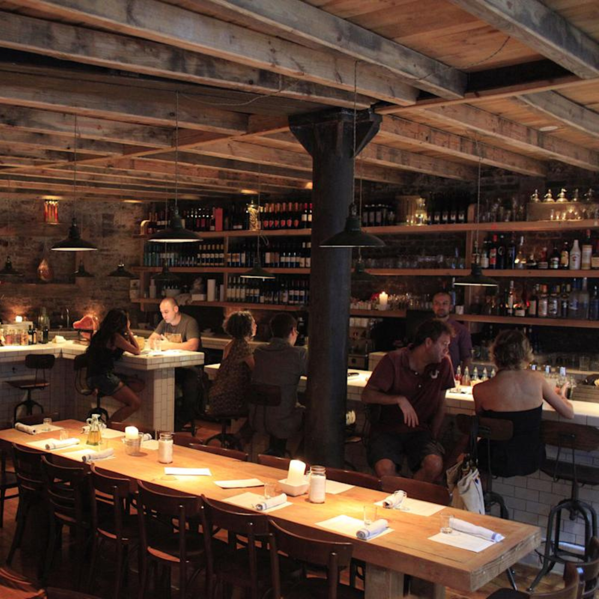 Rustic bar with wood beams, brick walls, warm lighting, and guests enjoying drinks at a cozy communal table and stocked counter