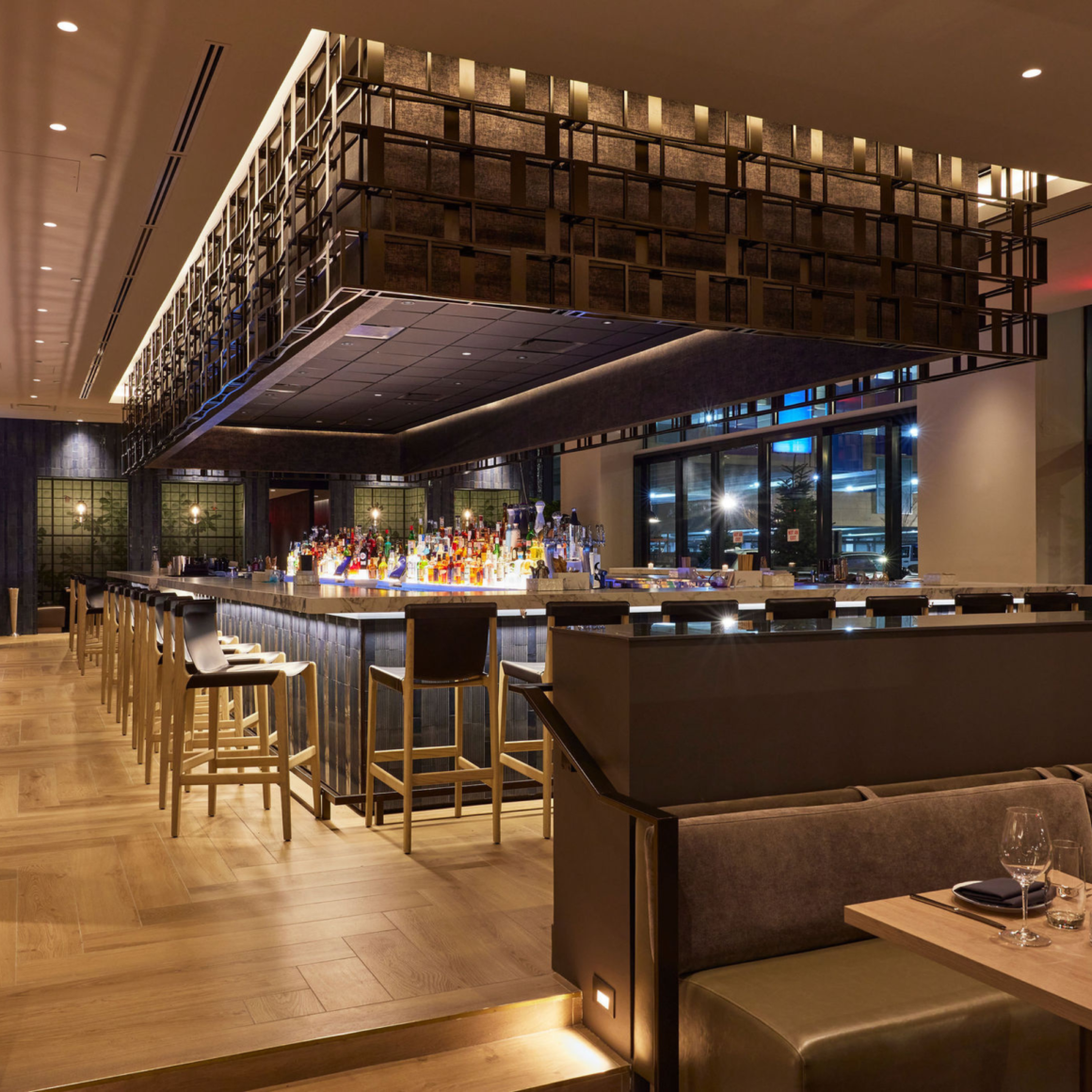 Elegant bar with high stools, geometric overhead decor, and backlit liquor display; cushioned booths and wine glasses set nearby