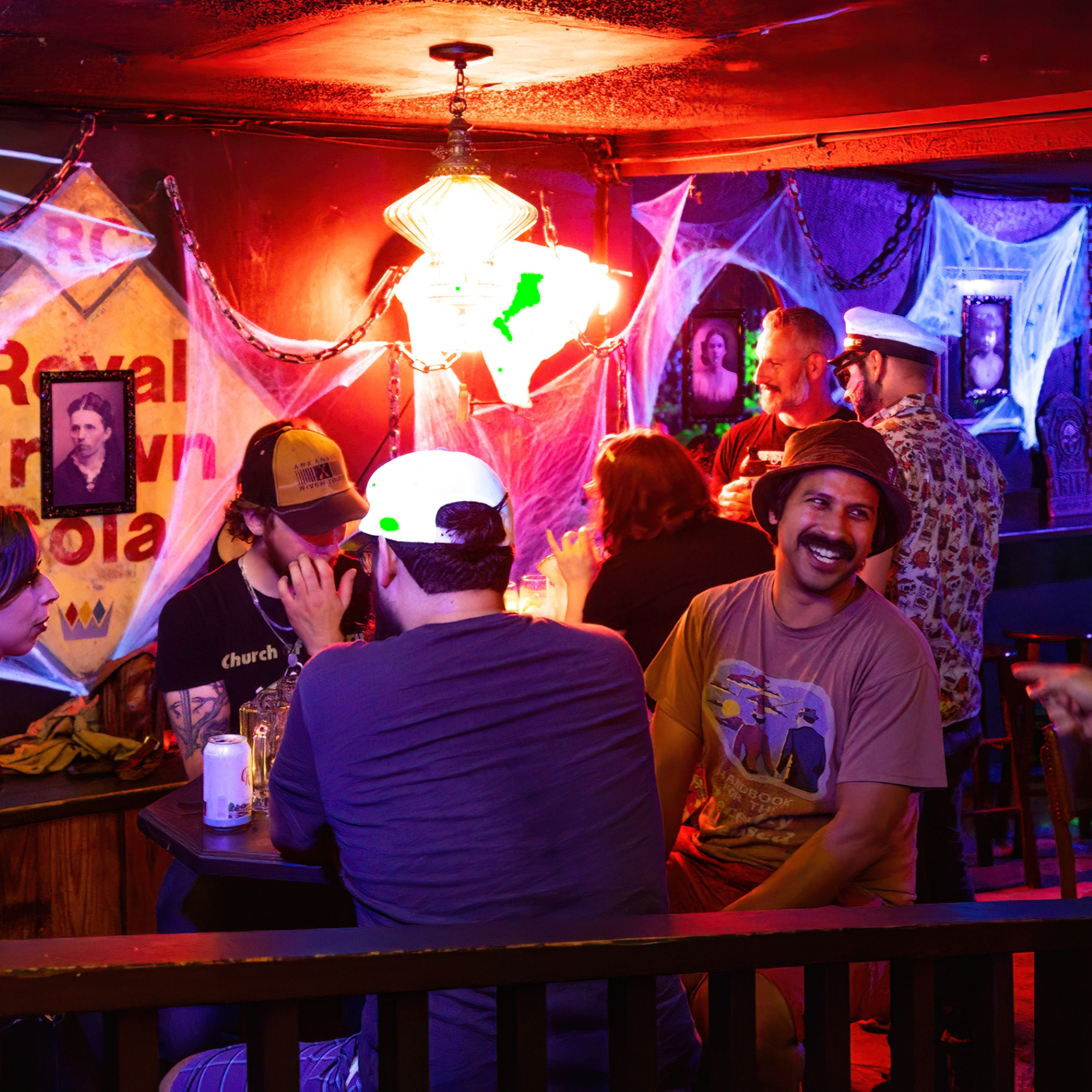 eople socializing in a dimly lit bar decorated with cobwebs, spooky lighting, and a “Royal Crown Cola” sign
