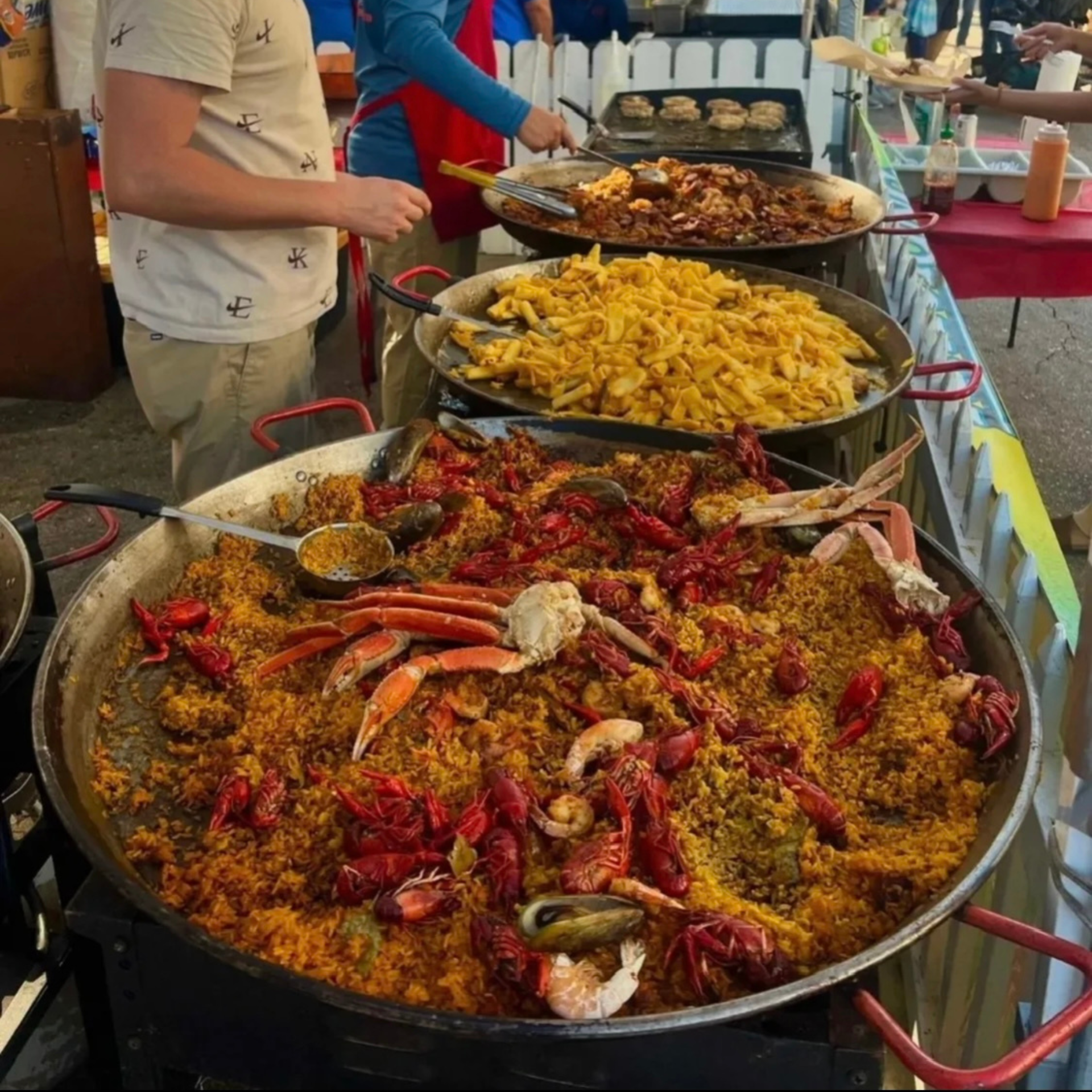 Seafood paella, creamy pasta, and grilled meats served in large pans at outdoor food stall