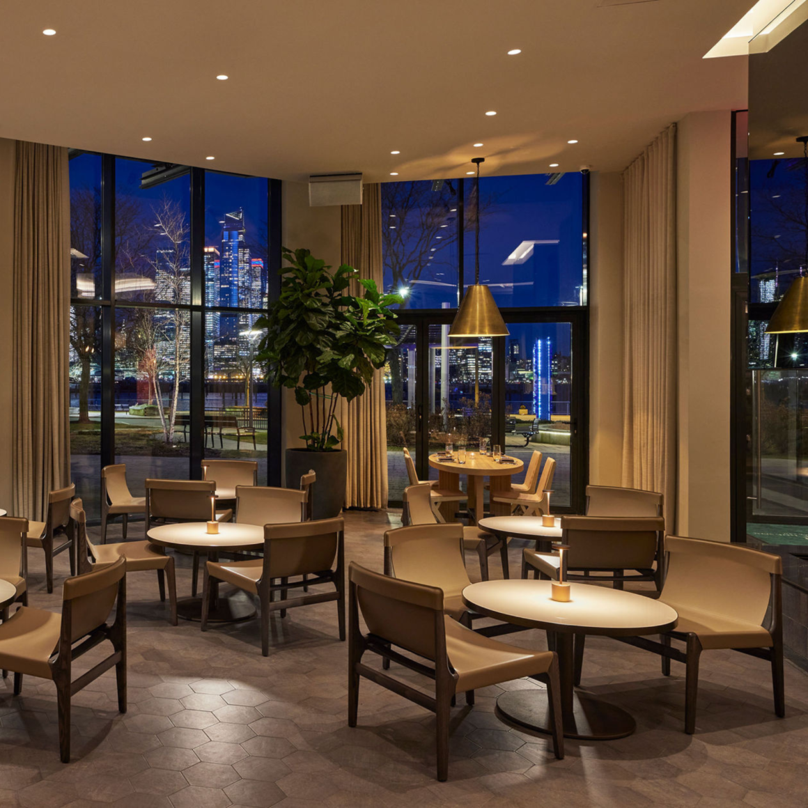 Upscale restaurant interior with city skyline view through floor-to-ceiling windows, warm lighting, round tables, and plush chairs