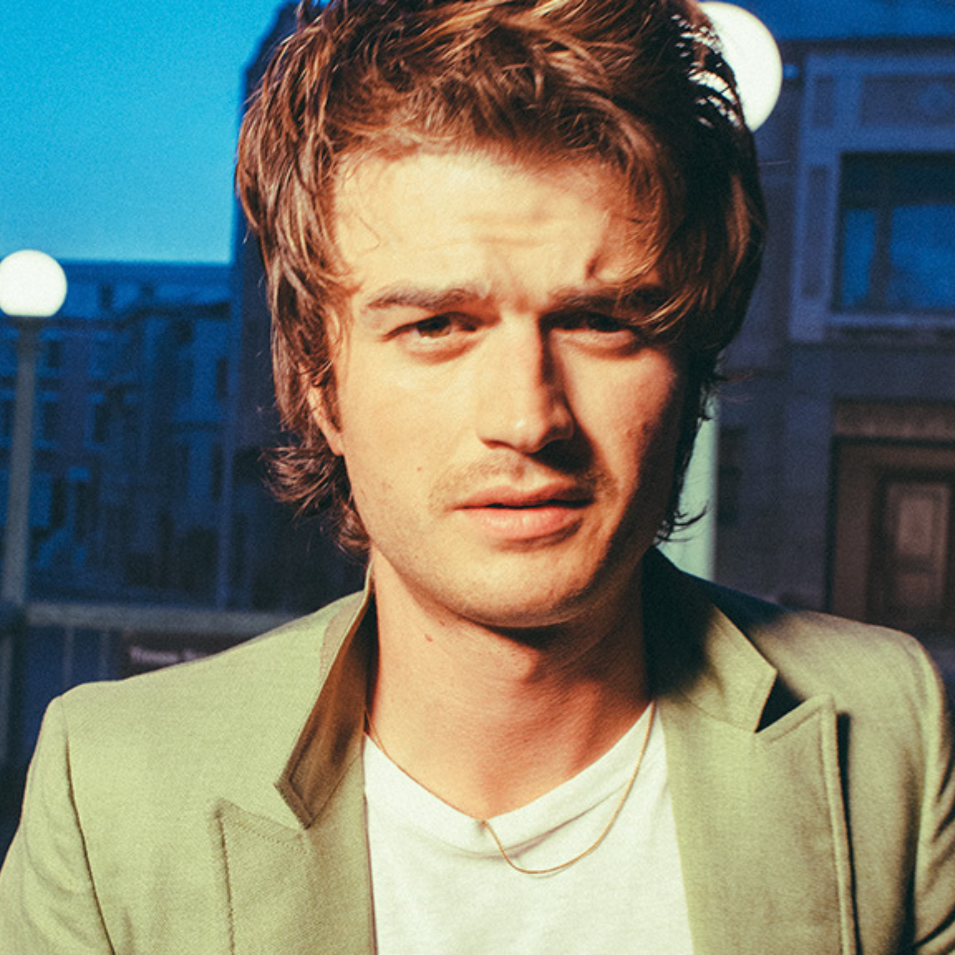 Djo (Joe Keery) in light green blazer and gold necklace, posing in an urban evening setting