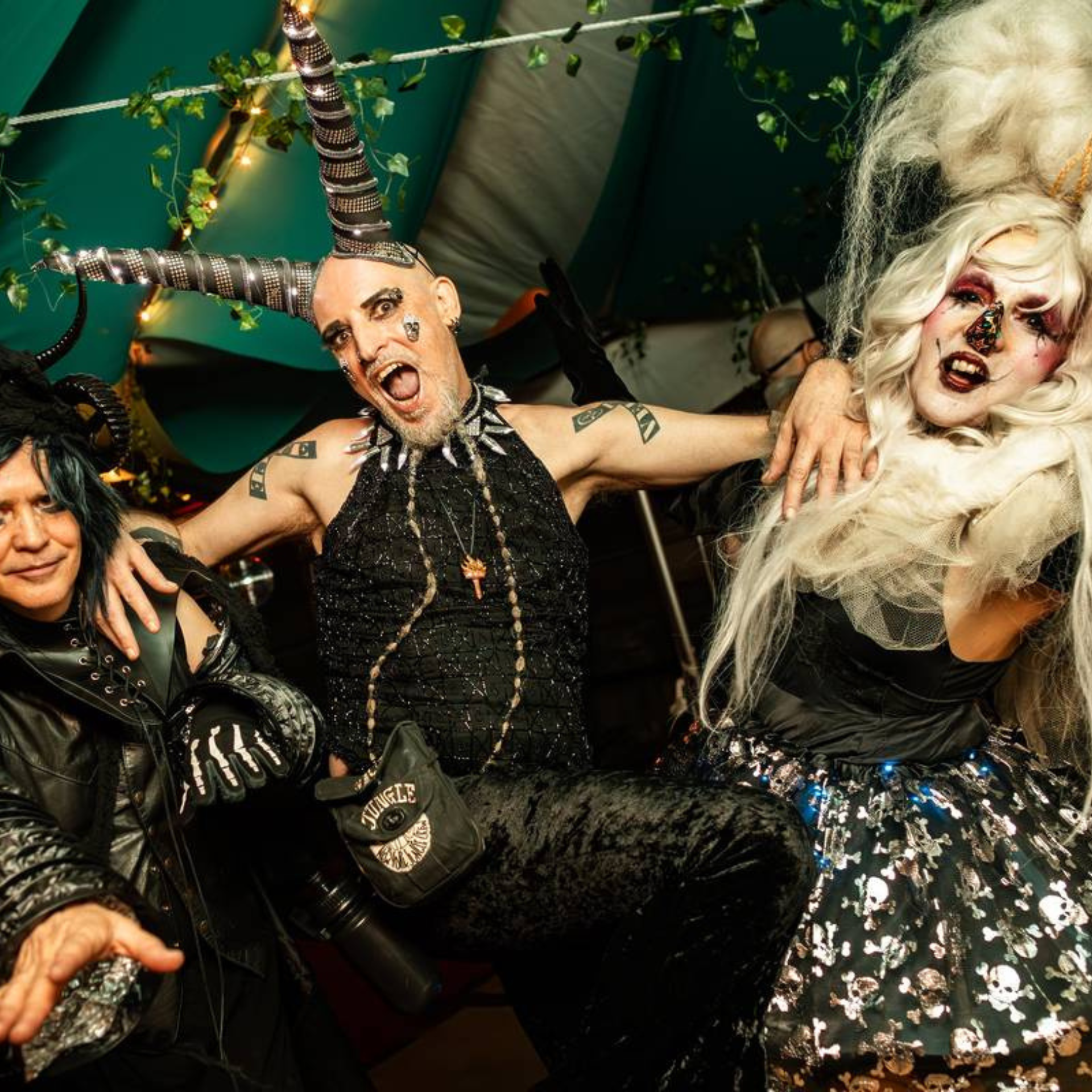 Three costumed guests pose at a festive event—horns, wigs, and dramatic makeup shine under string lights and lush decor