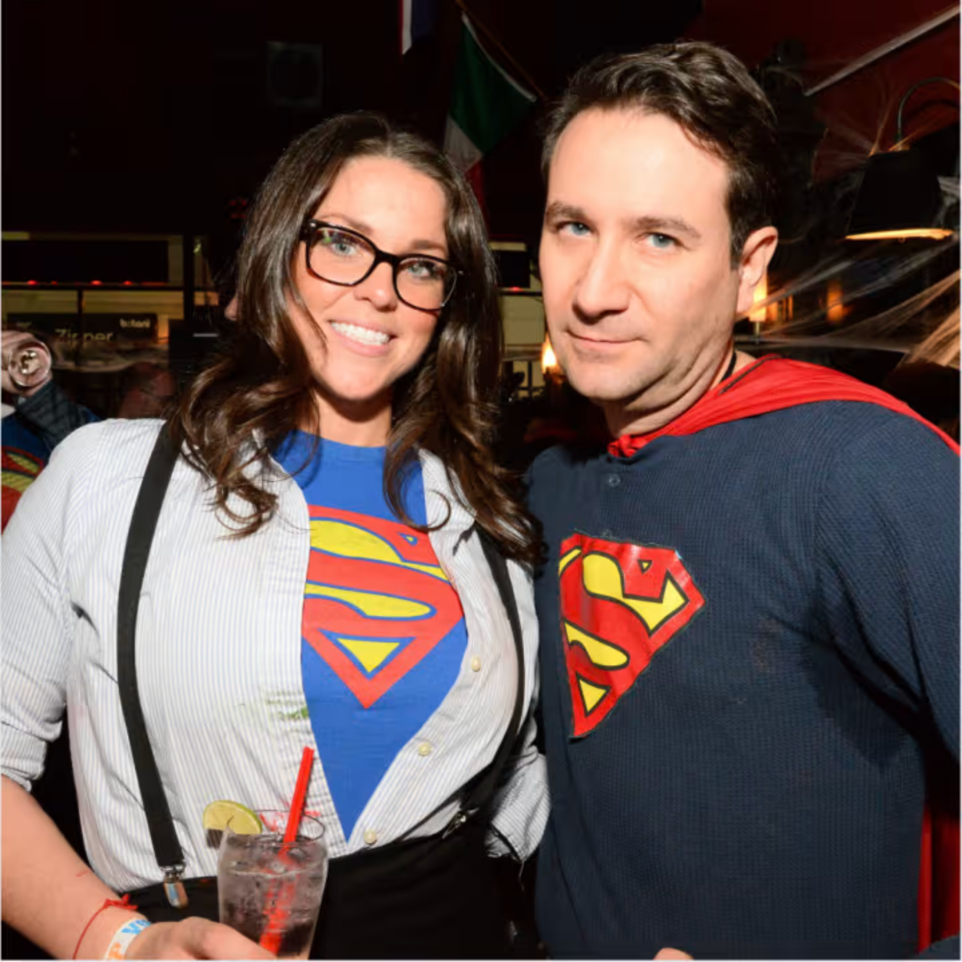 Two guests in Superman gear pose at a festive event with drinks, capes, and playful energy in a decorated party space
