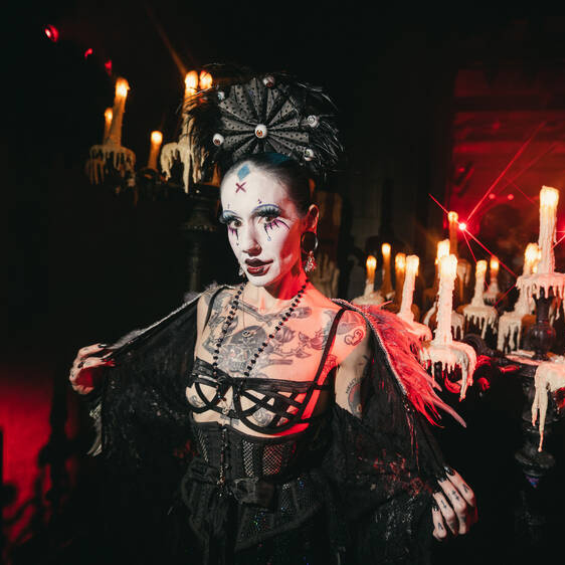 Person in elaborate gothic attire with dramatic makeup and black lace outfit, standing before candlelit candelabras in a dim, moody setting