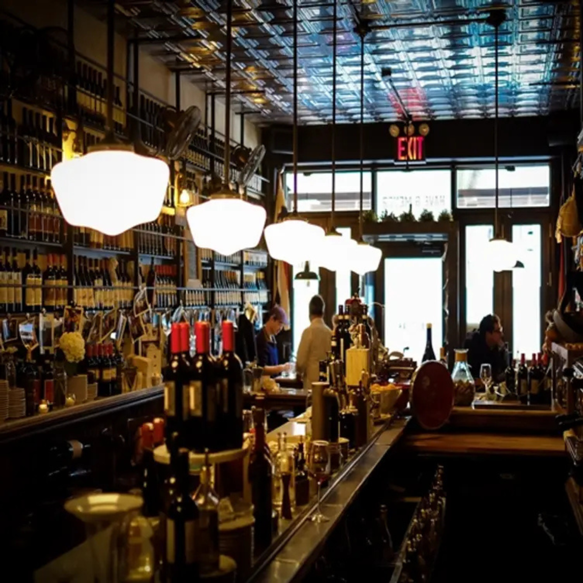 Vintage-style bar with tin ceiling, warm pendant lights, stocked shelves, and patrons enjoying drinks in a cozy, dim setting